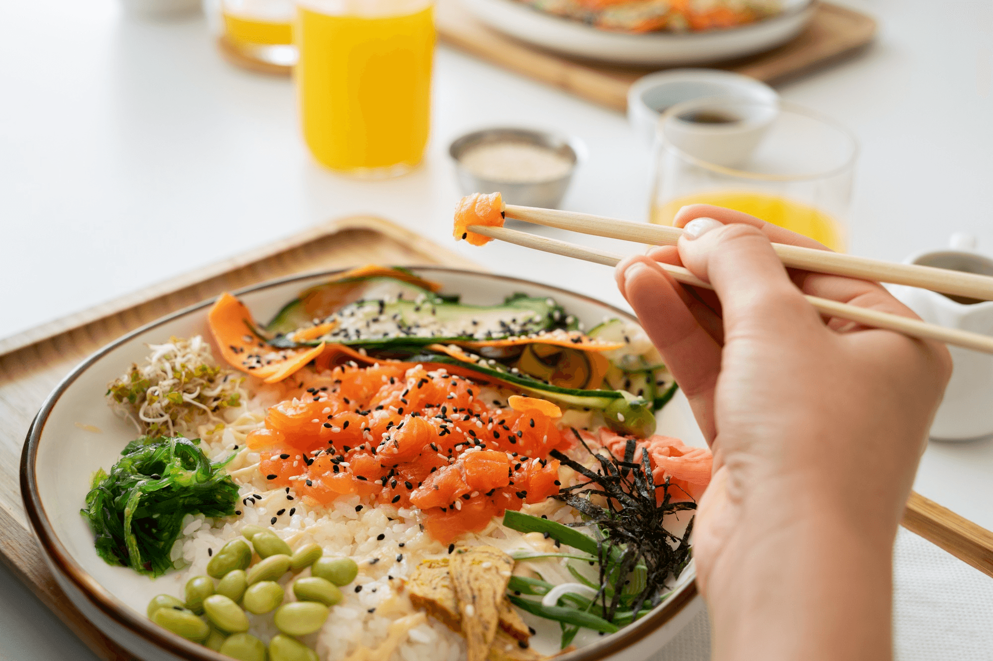A hand holding chopsticks picks up salmon from a colorful poke bowl with rice, vegetables, and seaweed. Bright and fresh, with orange juice in the background.