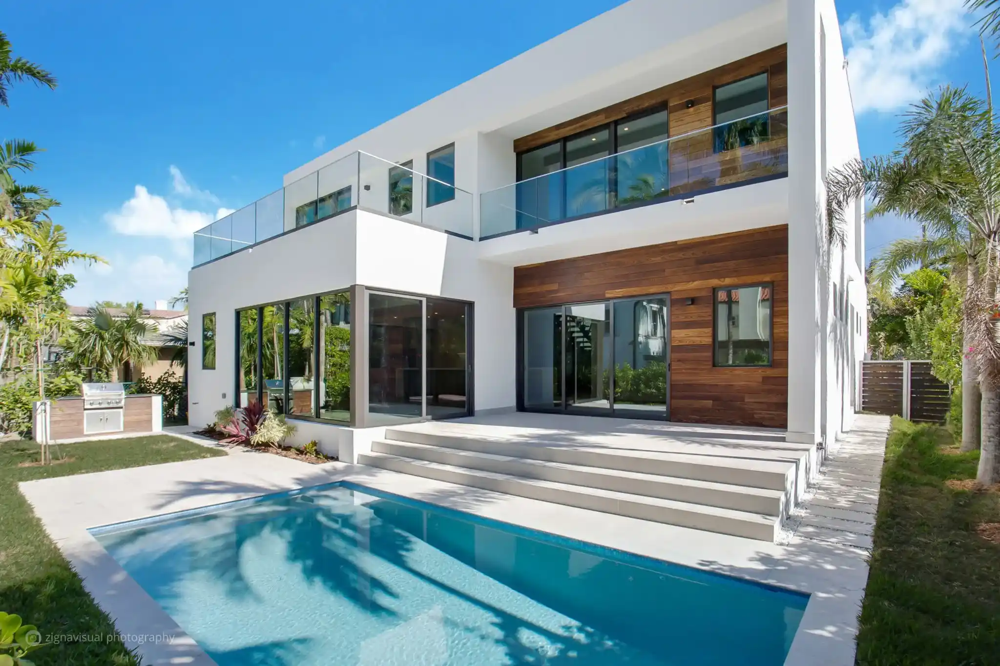 Modern Miami custom home by Atelier International with white stucco and wood-clad facade, glass-railed balcony, summer kitchen, and pool surrounded by tropical landscaping.
