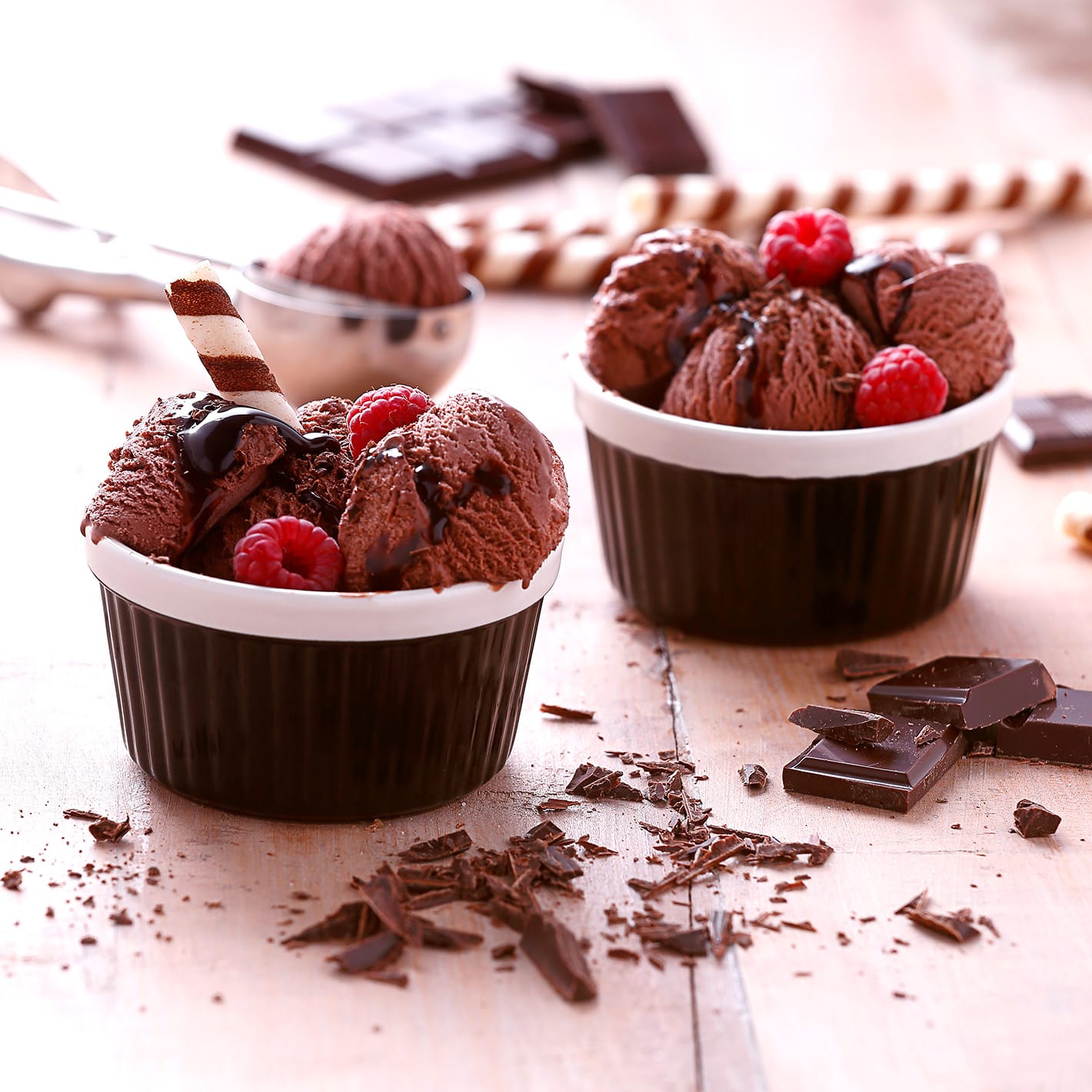 Two bowls of chocolate ice cream with raspberries, chocolate sauce, shavings, and wafer sticks on wooden table