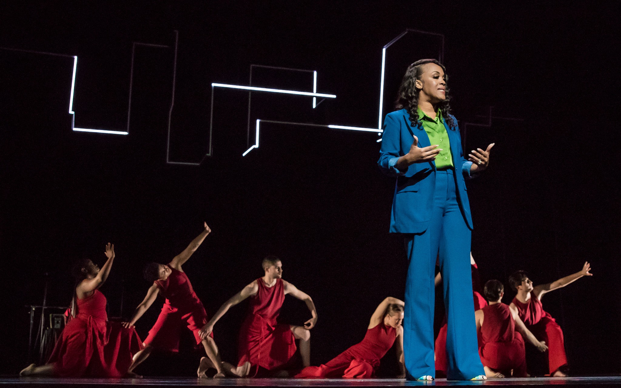 A performer in a blue suit speaks on stage, with dancers in red outfits behind, set against a dark background.