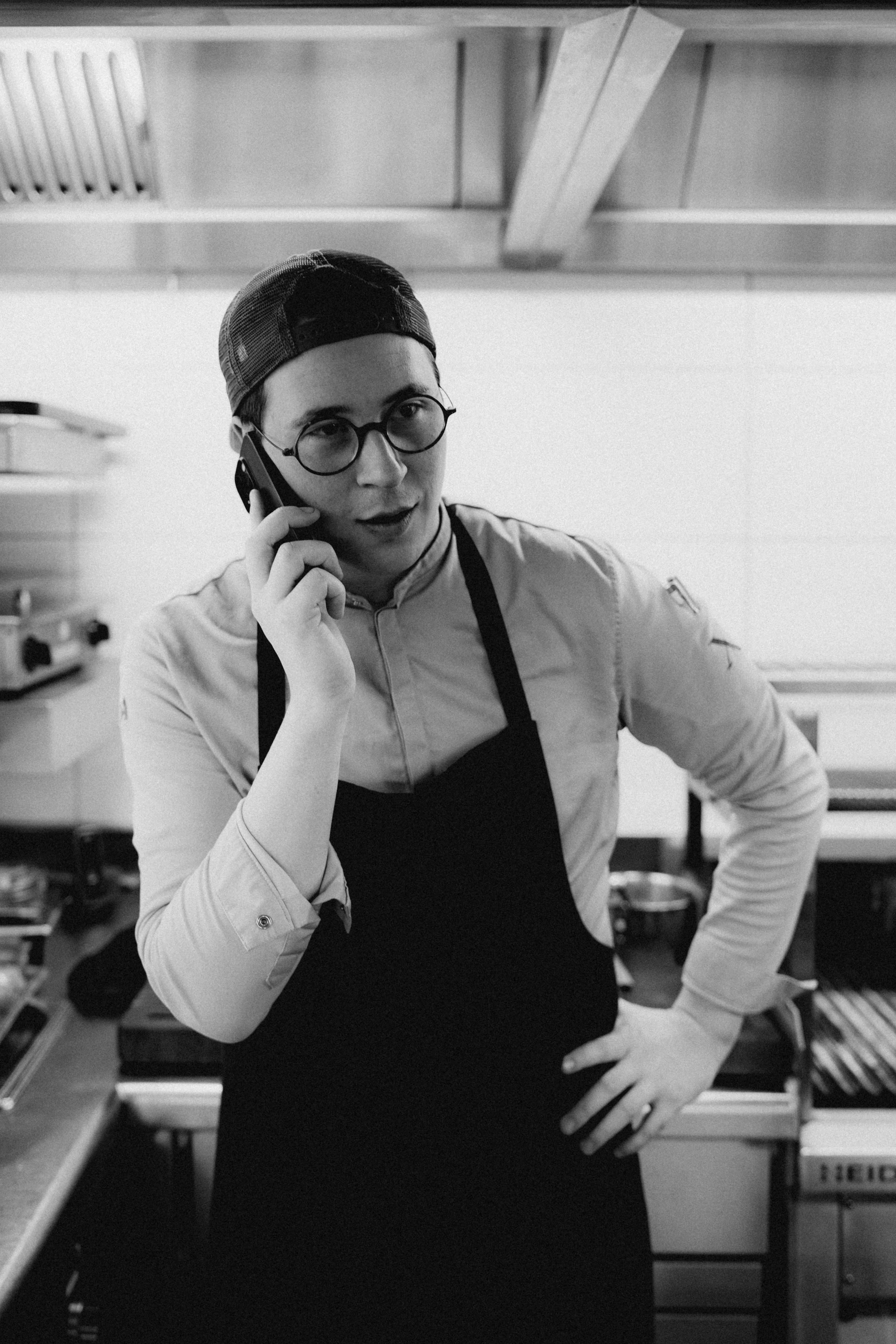 A person in an apron speaks on the phone while standing in a kitchen environment, focused and engaged.