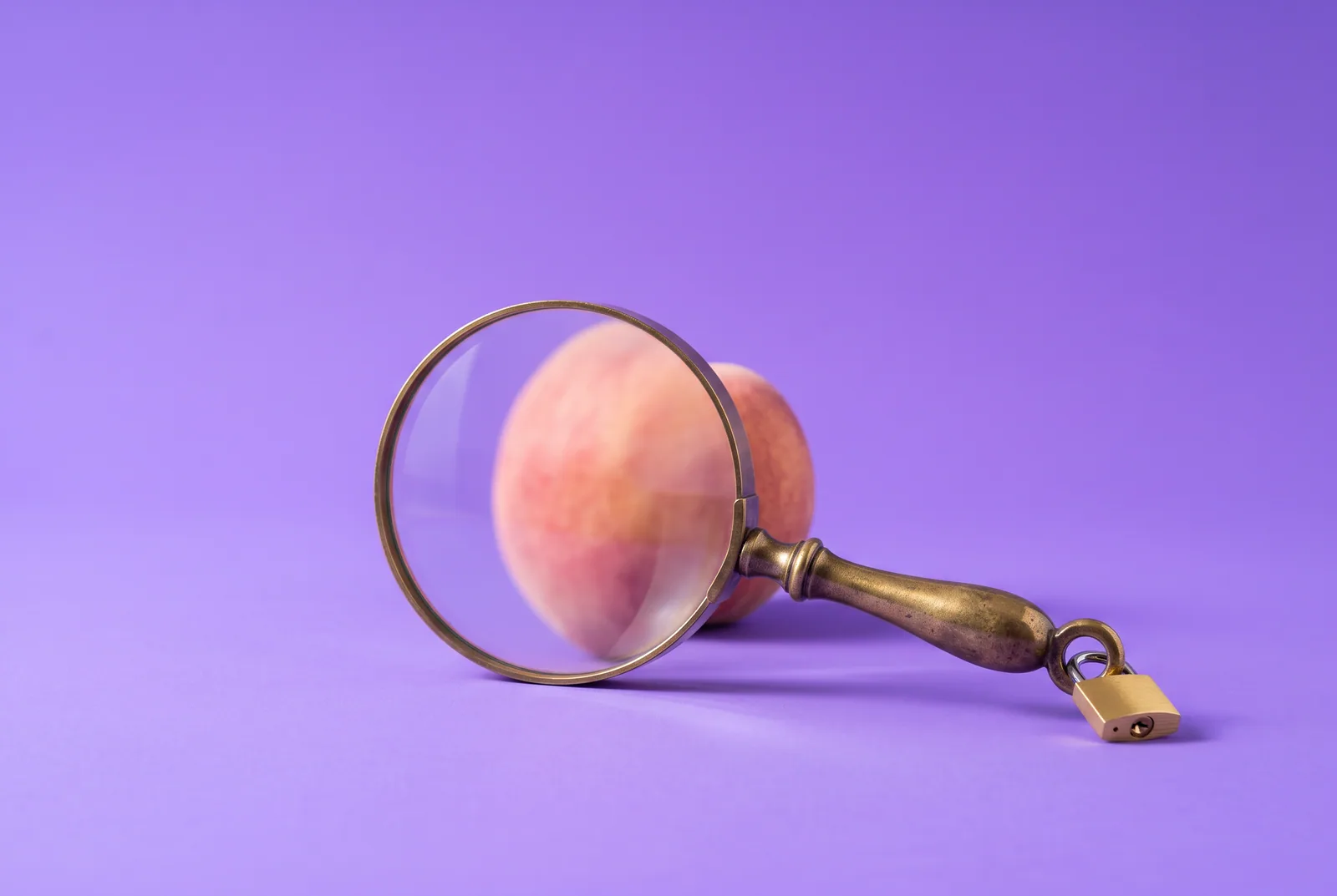 Brass magnifying glass with a small gold padlock on its handle in front of a blurred peach on a lilac backdrop, a metaphor for how to lock SafeSearch