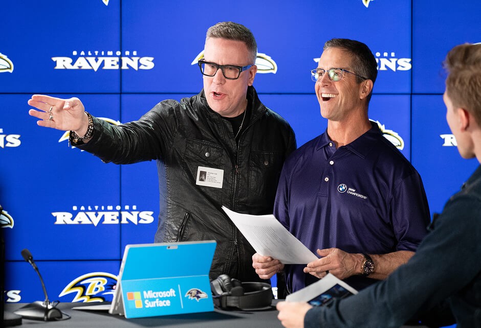 Two men stand in front of a Baltimore Ravens backdrop; one gestures while the other holds papers and smiles, with a Microsoft Surface on the table in front of them.