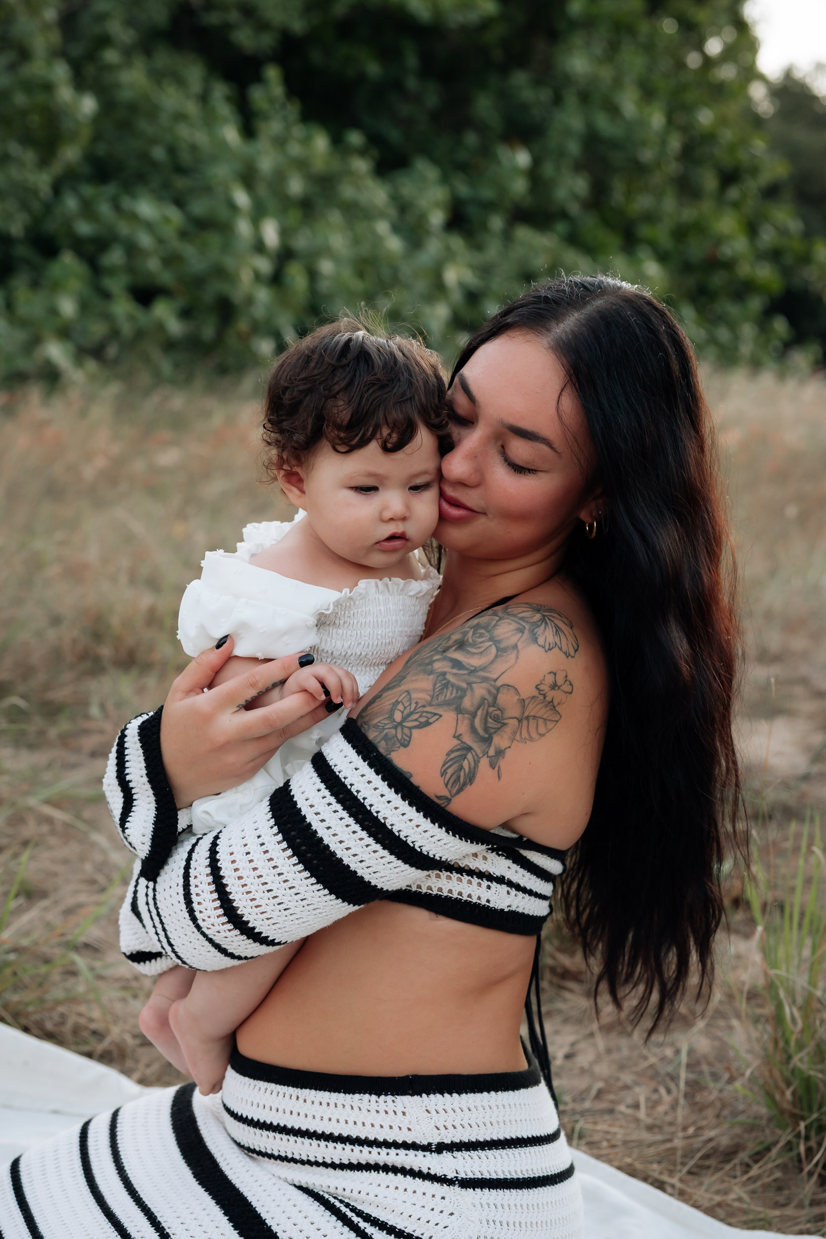 Mother and daughter sharing a cuddle in golden sunset light while standing in an open grassy field during a lifestyle motherhood photoshoot.