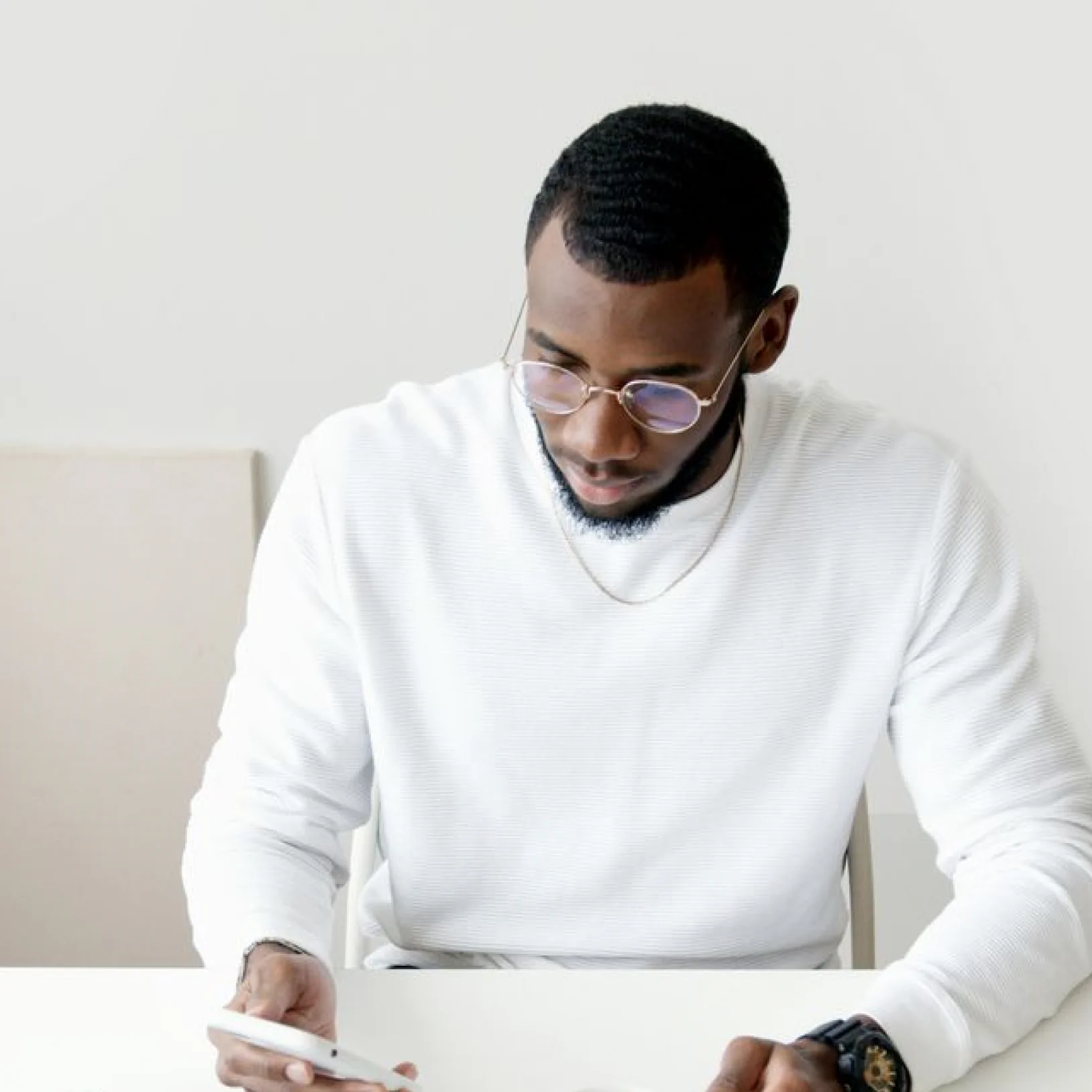 A man looking down intently while sitting at a desk against a minimalist white background. He has a neatly trimmed short beard, wears round metallic-framed glasses, and is dressed in a white long-sleeved textured sweater. His right hand holds a white smartphone or tablet device on the desk surface, and he wears a black sports watch on his left wrist.
