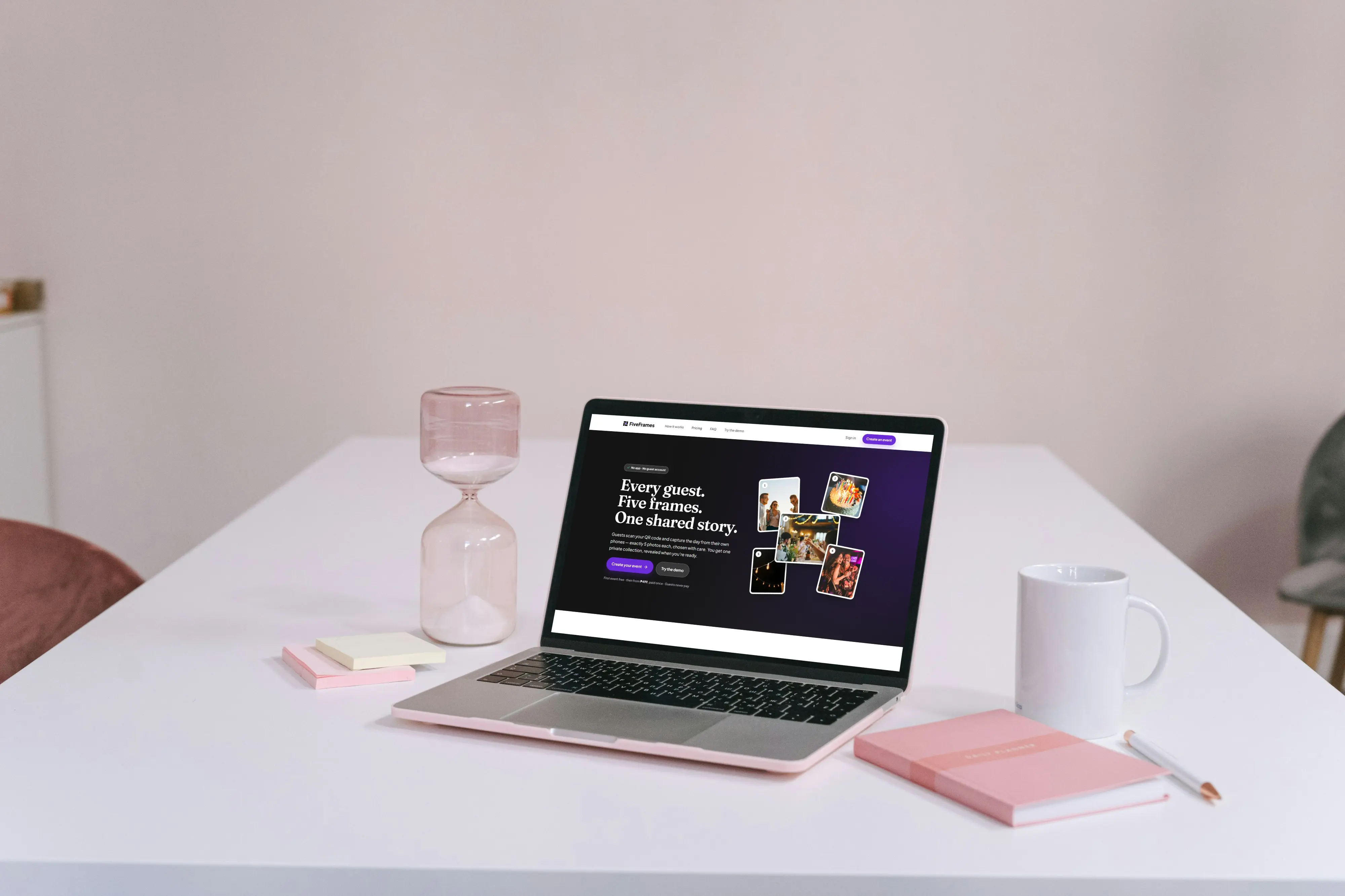 Laptop on a white desk beside a pink hourglass, sticky notes, a white mug and a pink notebook, showing the FiveFrames homepage with the headline “Every guest. Five frames. One shared story.” and a collage of event photos.