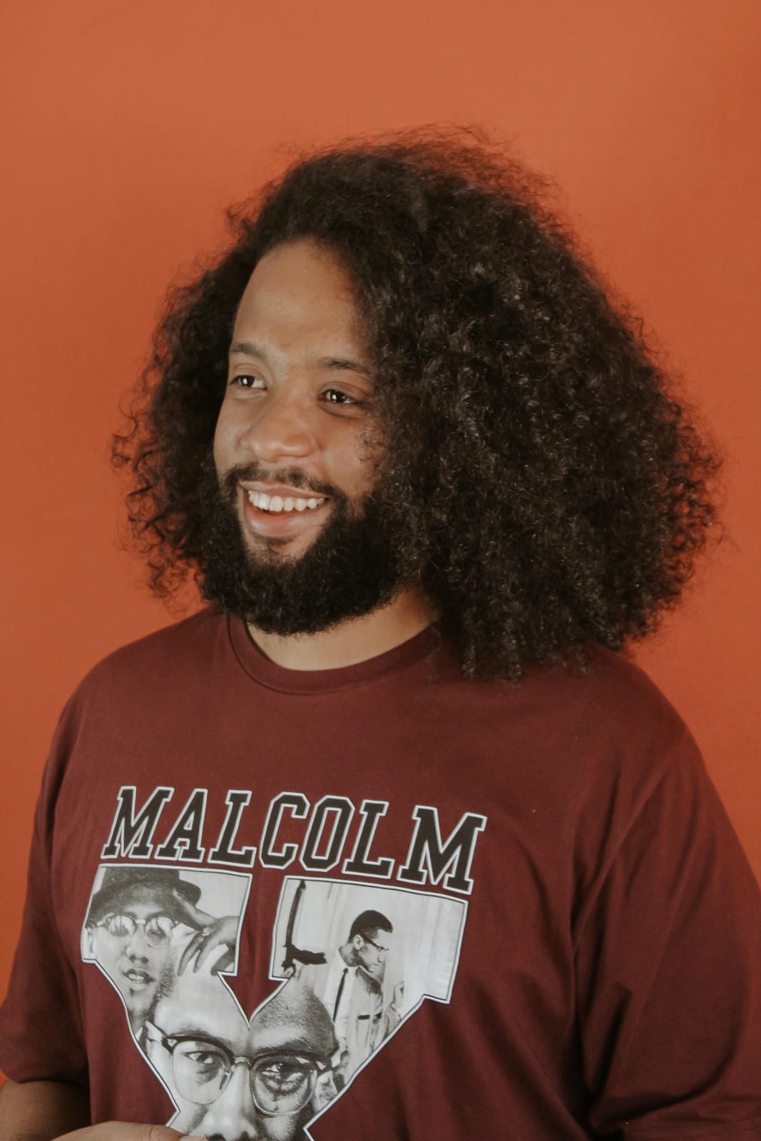 Homem negro sorrindo, cabelo comprido e barba, camiseta vinho no funo learanja