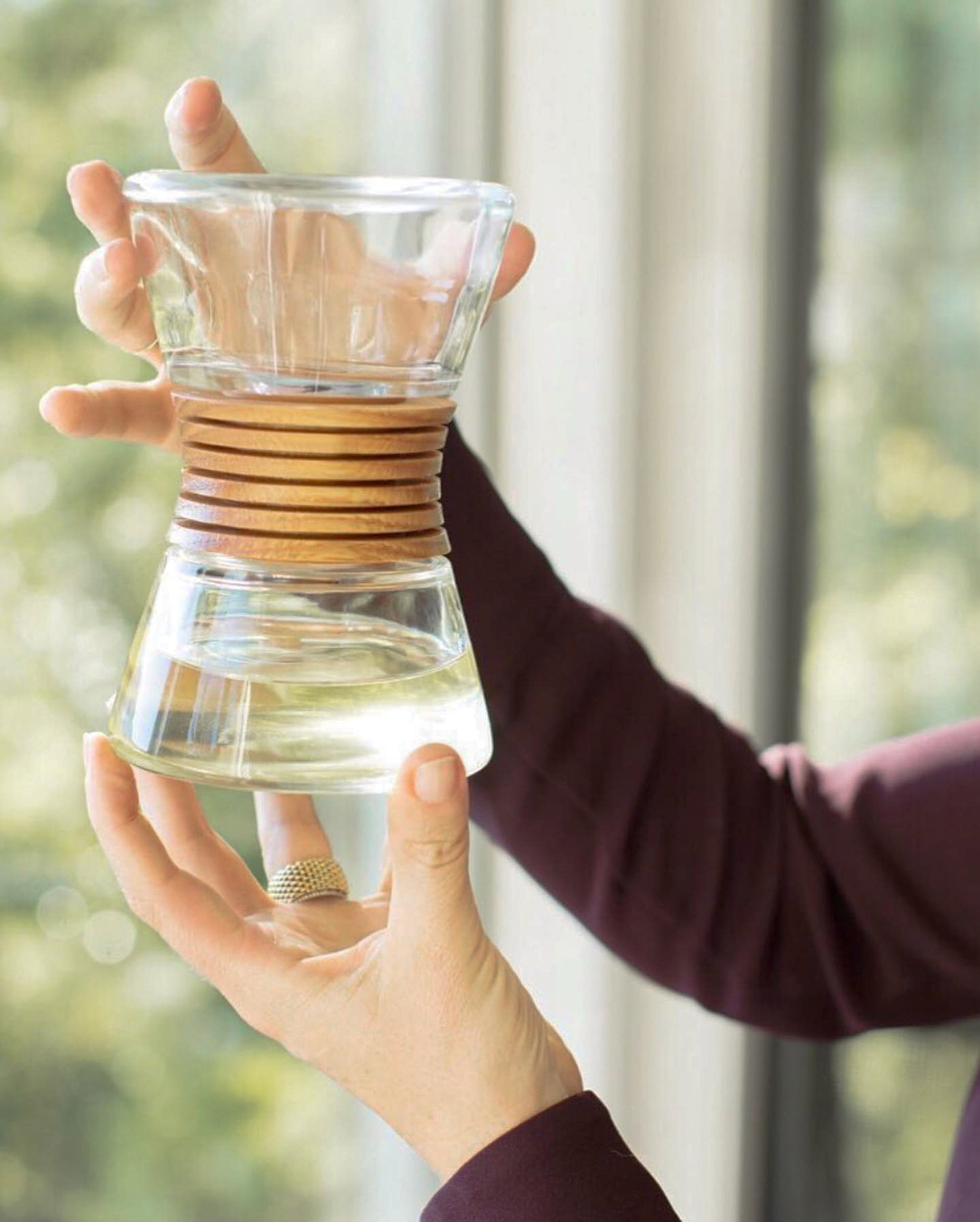 Hands holding hour-shaped glass bottle with wooden diffuser in the middle.