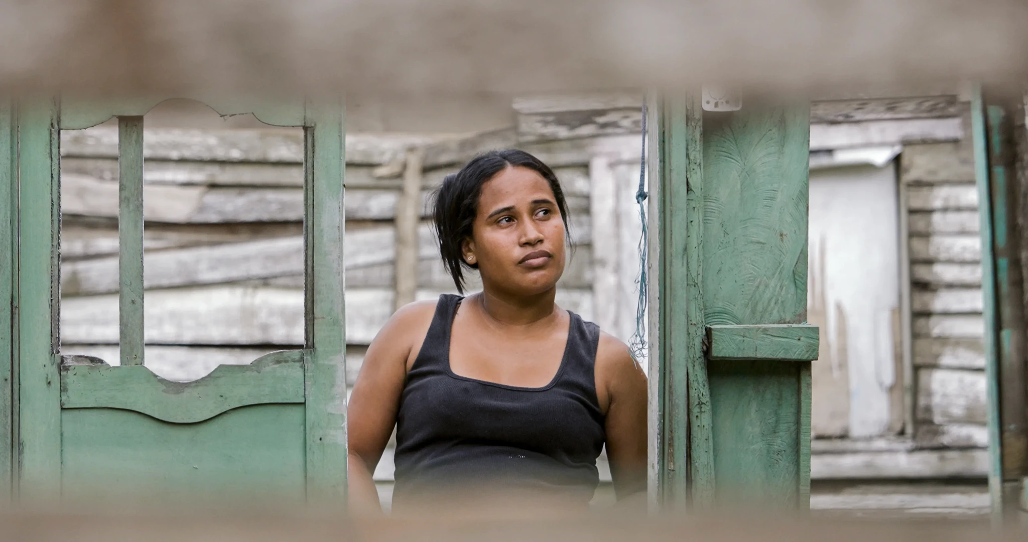 A woman in a black tank top stands in a rustic, wooden doorway, looking thoughtfully into the distance. The weathered wooden walls and doors add a rustic feel to the scene.