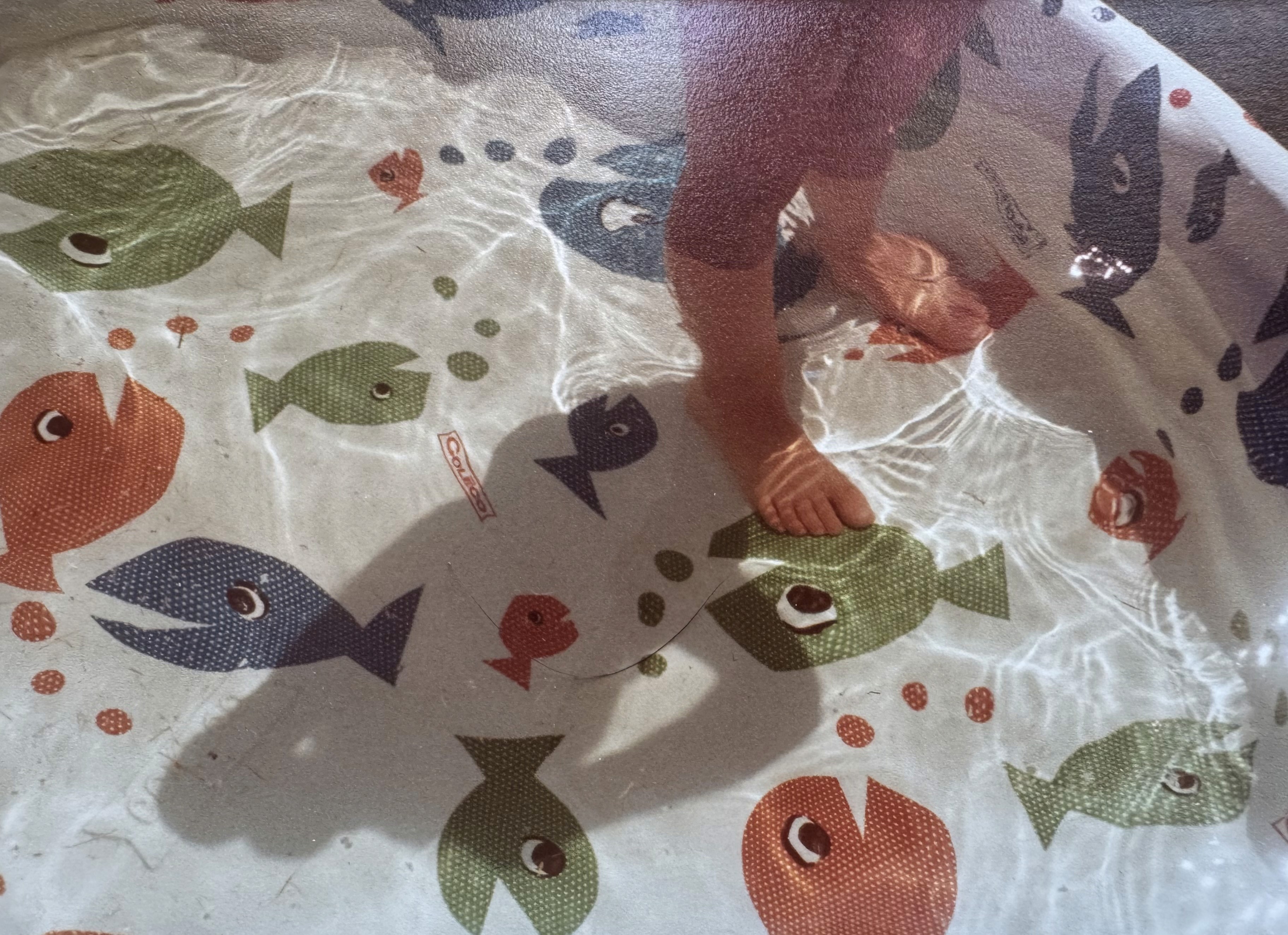 A little girl's feet standing in a kiddie pool that's filled with water and has a design of blue, green, and orange fish on a white background. 