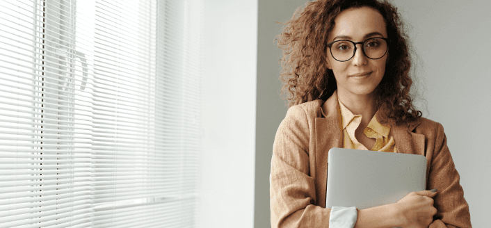 Woman with glasses holding laptop in office setting.