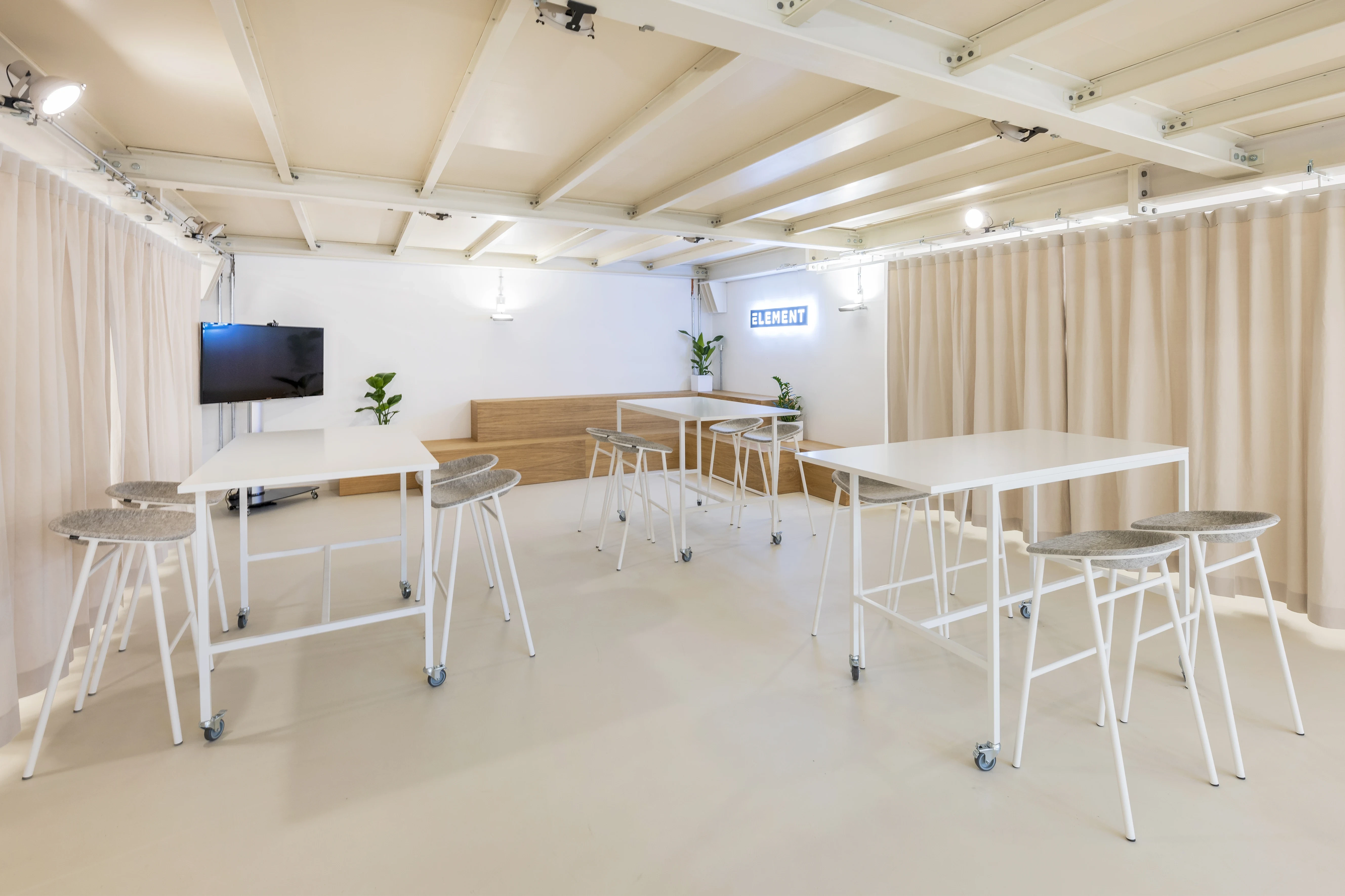 Workshop room at ELEMENT with white standing tables on castors, grey upholstered bar stools, beige curtains on both sides, a wall-mounted TV, wooden bench and an exposed white beam ceiling.