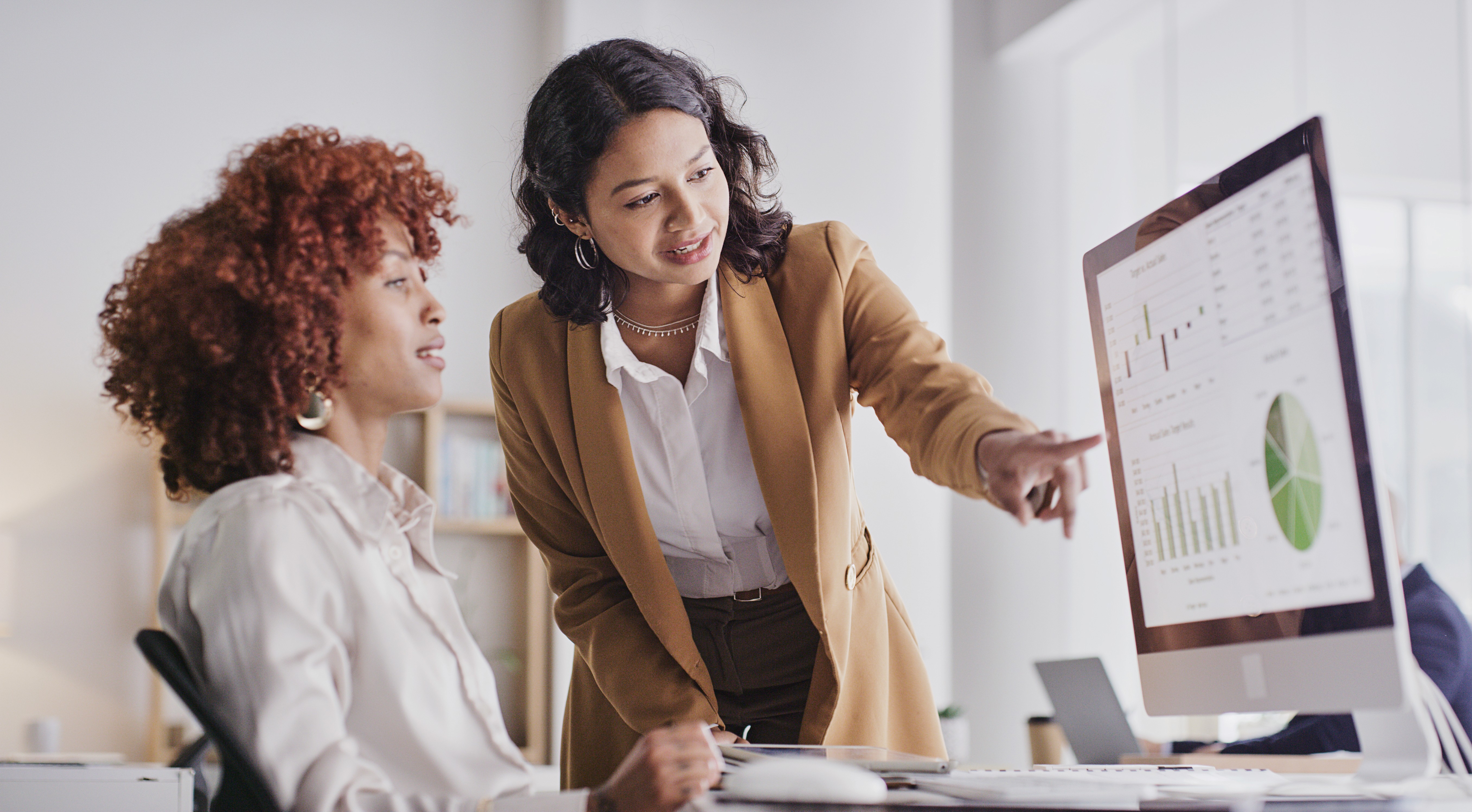 Two women discussing information while looking at a computer screen in a bright office setting.