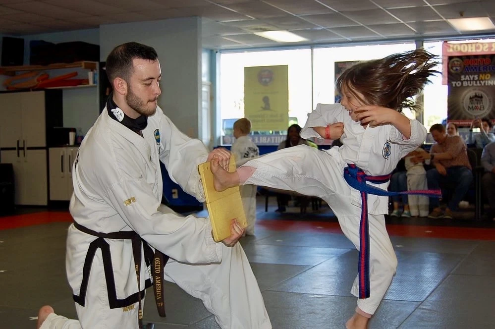 Instructor holding a board while a student breaks it with a jumping side kick.