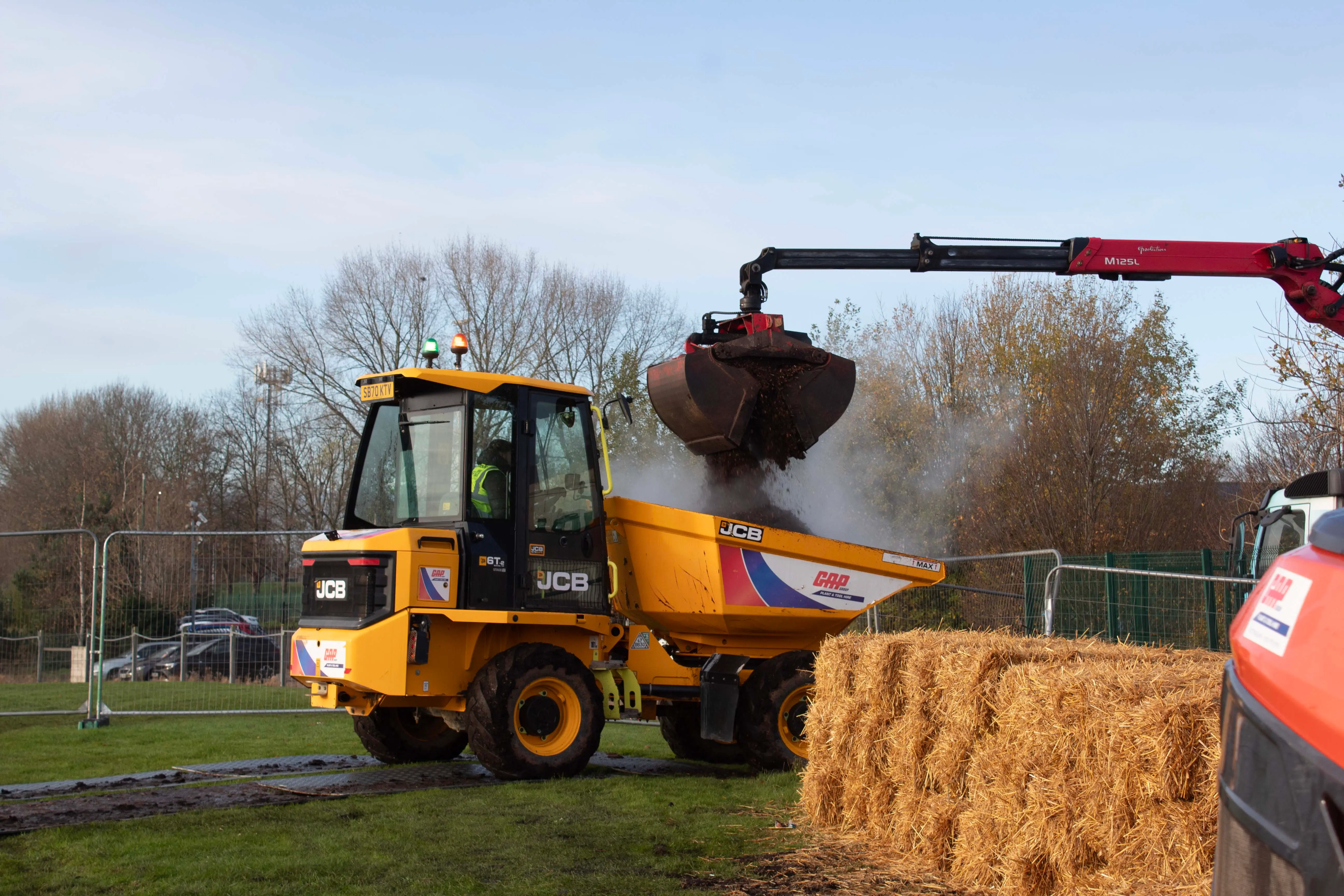 A yellow construction vehicle lifts a load while parked near stacked hay bales in a field on a sunny day.