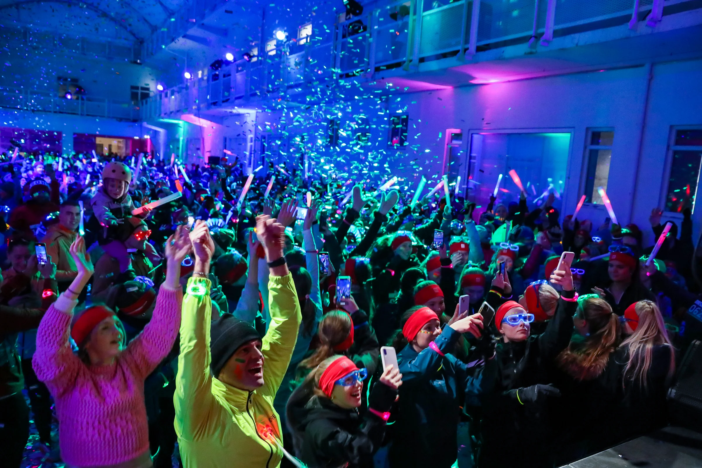 A large crowd celebrates at the pre-Northern Lights Run party in Reykjavík, raising glowing coloured foam sticks and phones beneath falling confetti and blue and green stage lighting.