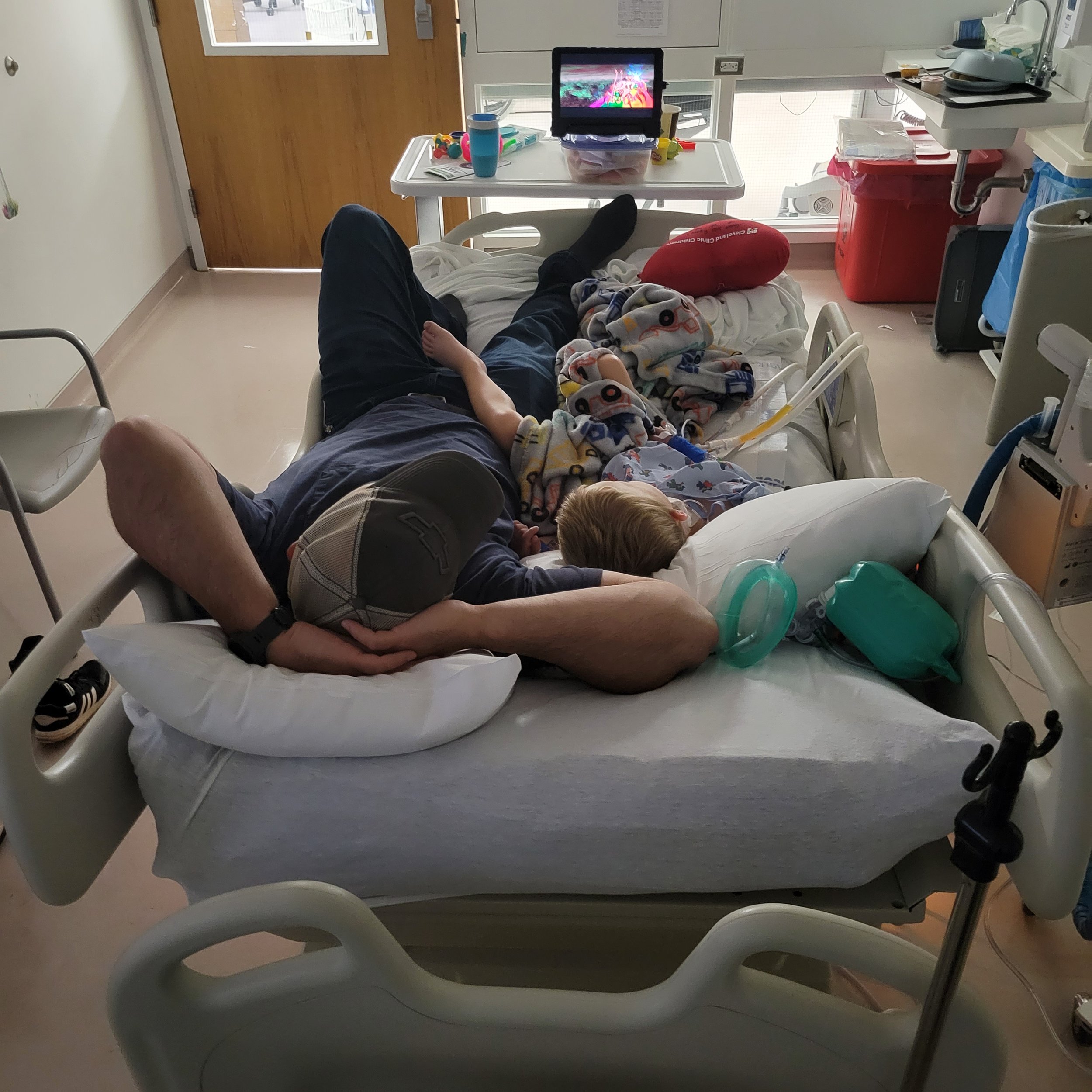 father and son lay in hospital bed in pediatric intensive care unit watching a movie on a tablet