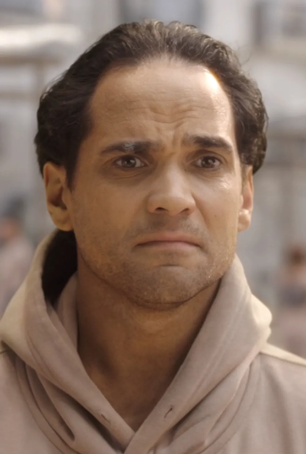 A contemplative man in a beige hoodie stands outdoors, expressing concern, with soft urban architecture blurred in the background.