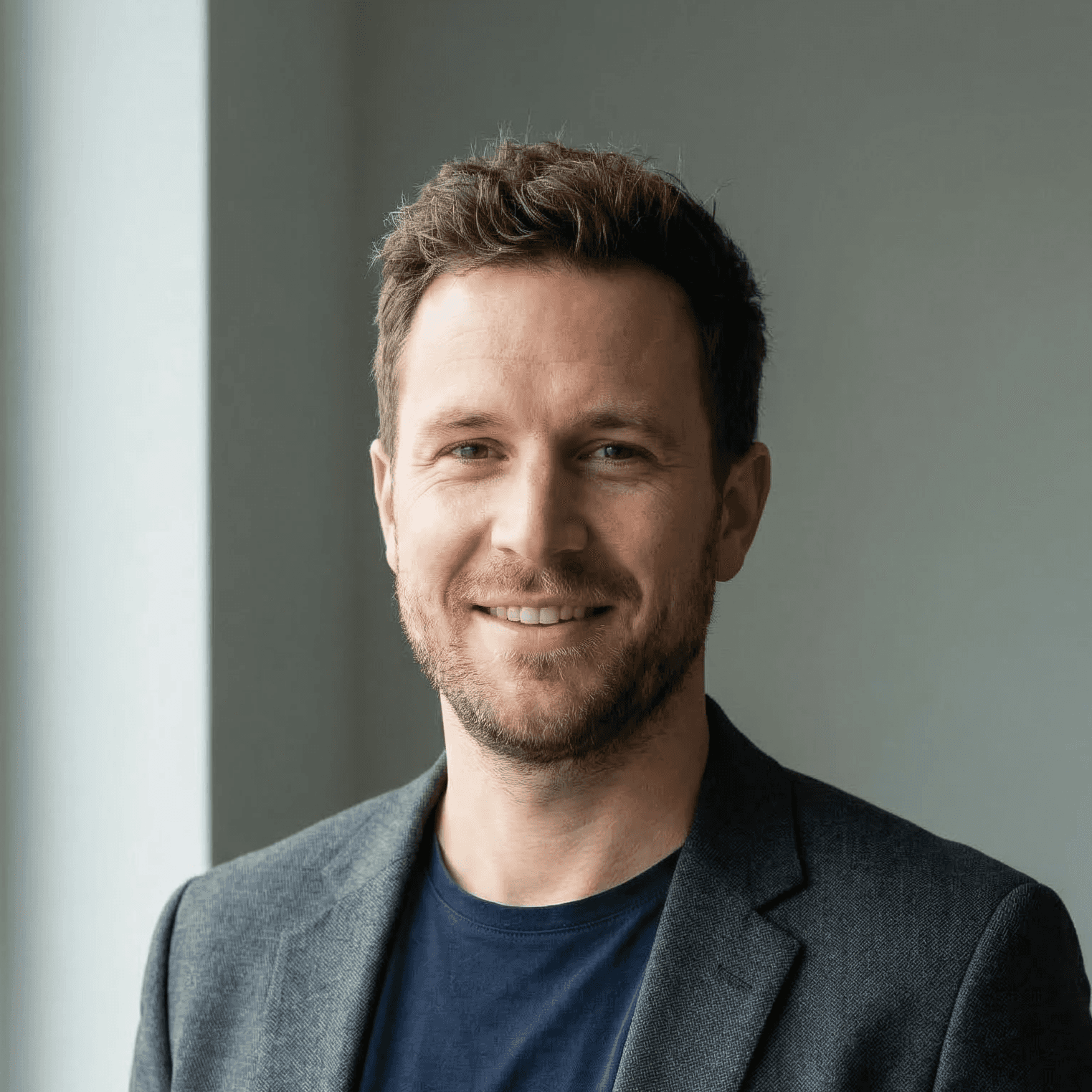 Smiling man with short brown hair and beard, wearing a gray blazer and blue t-shirt against a neutral wall.