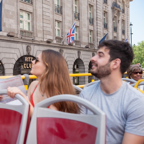 Twee mensen op een dubbeldekker-tourbus, met op de achtergrond het gebouw van de Ritz Club en de Union Jack-vlag.