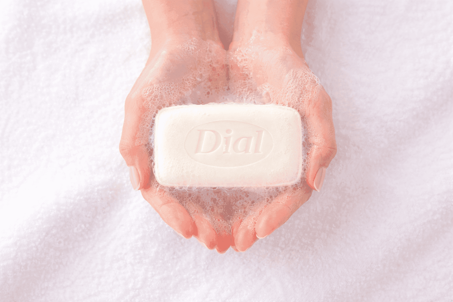 A bar of white Dial soap, lathery with foam, held by manicured hands against a background of soft, white towel.