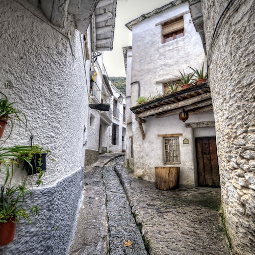 狭窄的鹅卵石小巷,两旁是粉刷成白色的建筑,有盆栽植物和一扇木门,门上方是一个乡村风格的阳台。