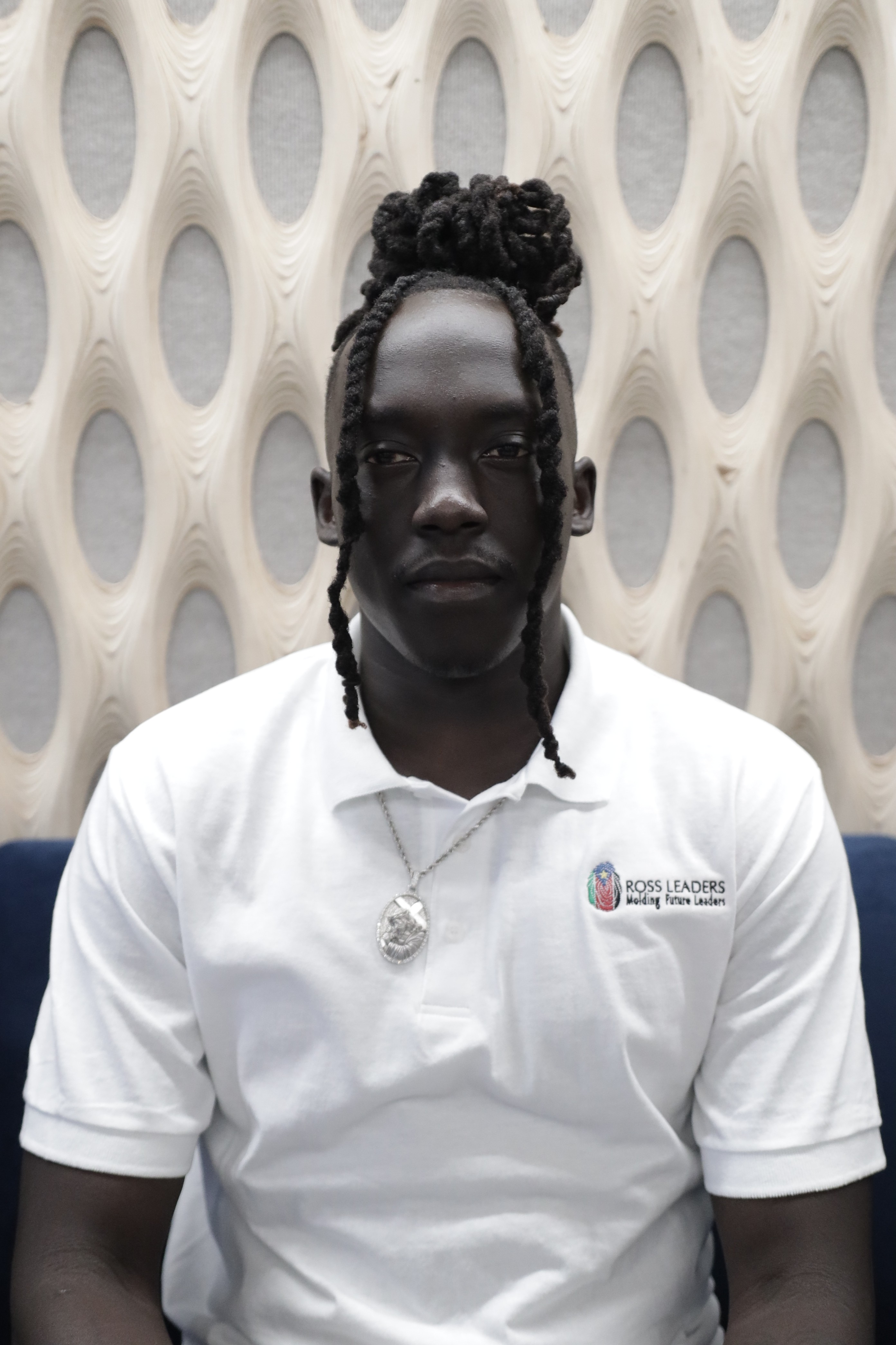 Portrait of a person with red hair, wearing a rust-colored outfit and scarf, set against a neutral gray background.