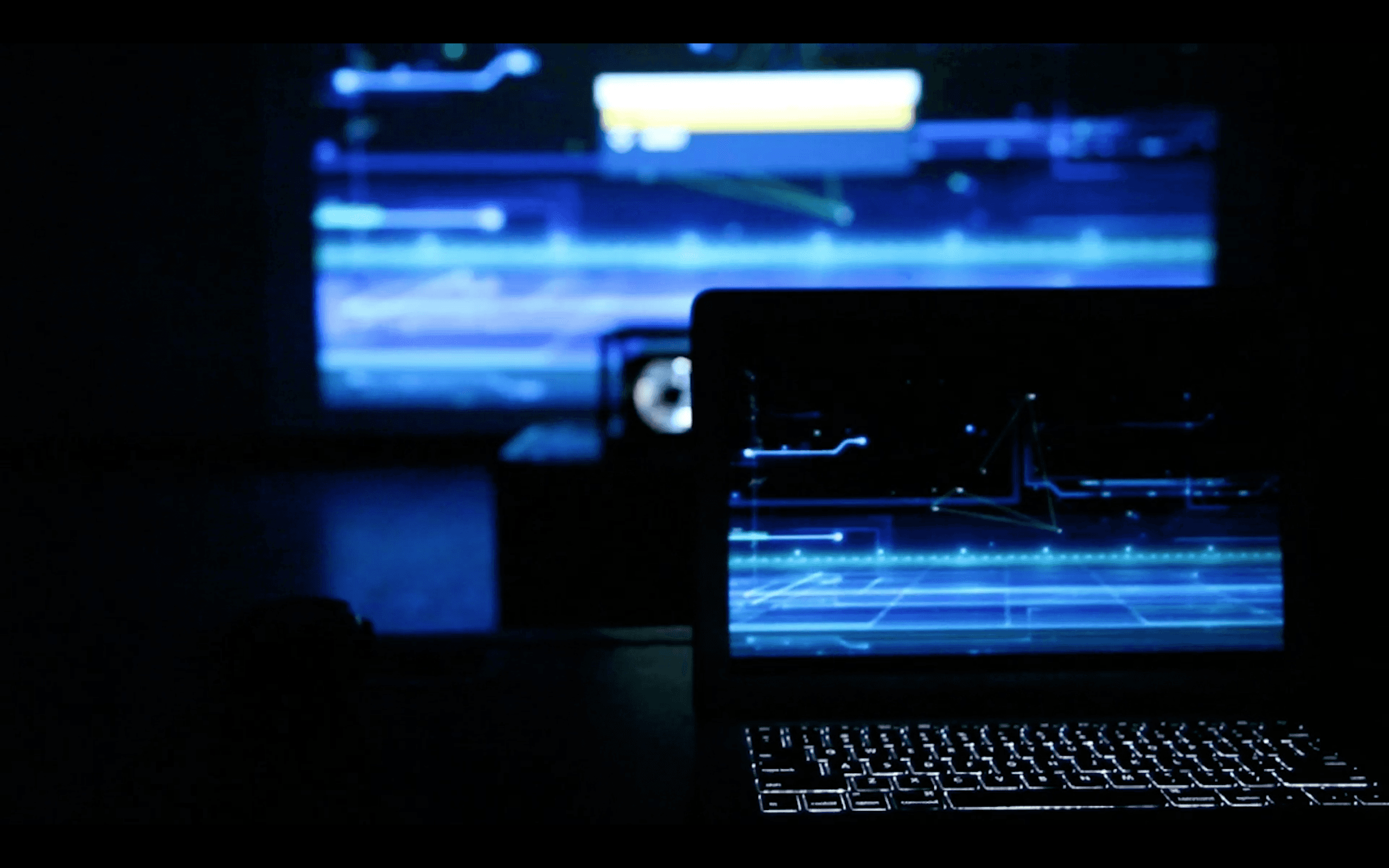 A dimly lit room features a glowing laptop on a table, with a digital interface displayed on its screen, mirroring the high-tech, futuristic graphics projected on a large screen in the background, conveying a theme of technology and innovation.