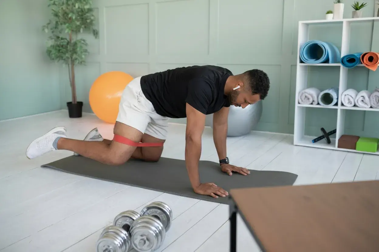A man exercises with a resistance band on a yoga mat in a modern home gym, with dumbbells, a stability ball, and neatly stacked yoga mats on shelves nearby.