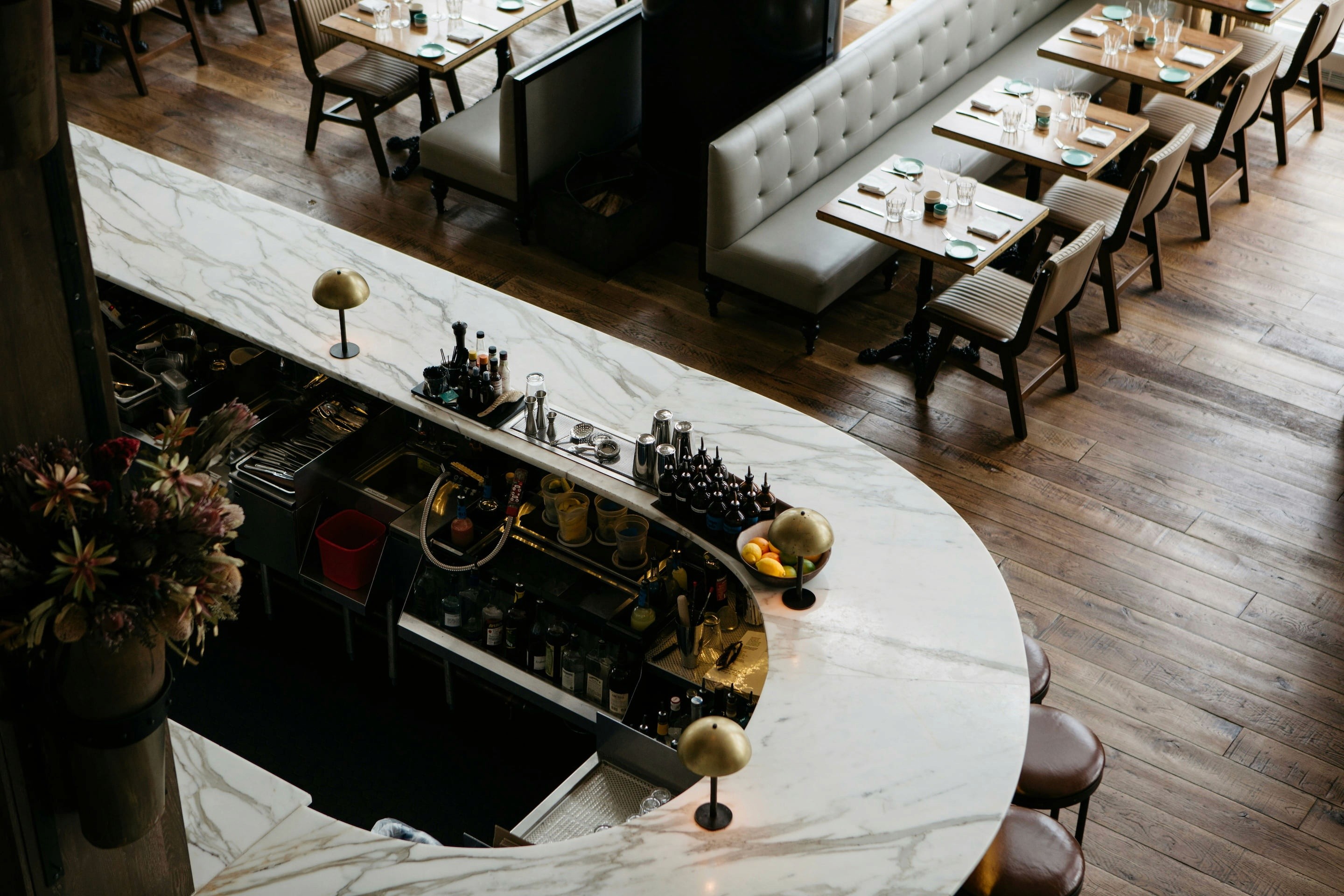 Aerial view of a stylish restaurant with a marble-topped bar, wooden floor, and beige cushioned booth seating. Tables set elegantly, evoking a cozy, upscale ambiance.