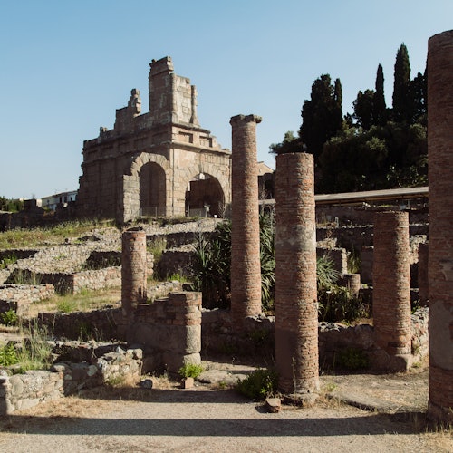 Ruines antiques avec des colonnes en brique et une arche en pierre sur un fond d'arbres et de ciel dégagé.