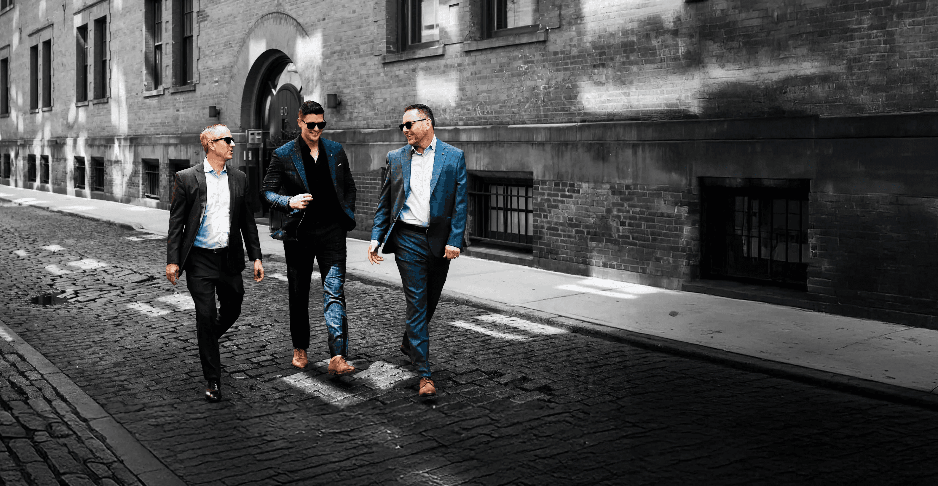 Three business professionals walk and talk along a cobblestone street beside a large building, dressed in smart casual attire.