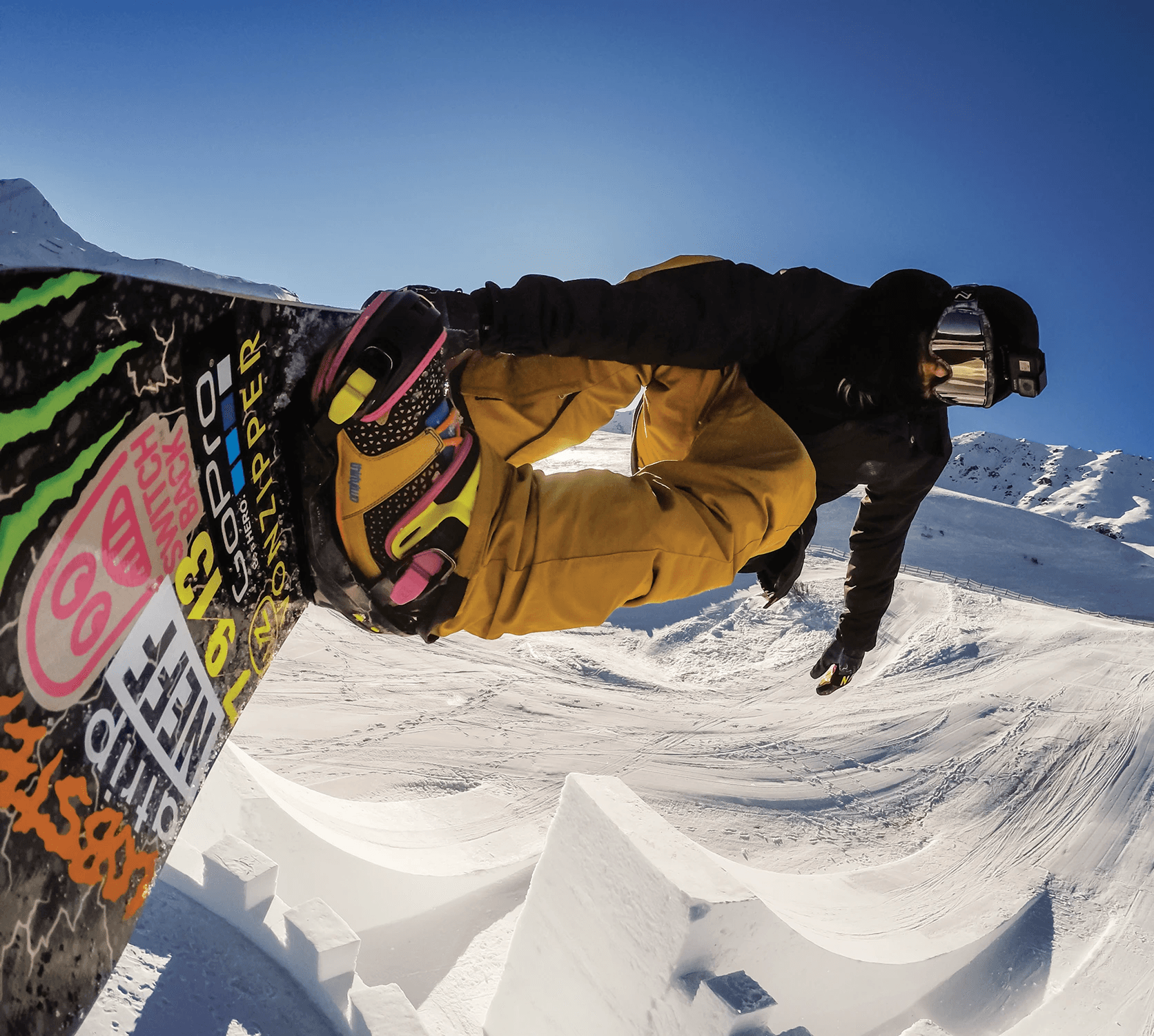 A GoPro camera mounted to the well-stickered board of professional snowboarder Halldor Helgason as he flies over a massive kicker built on a snow-covered Watles Ski Area in the Italian Alps. Halldor is wearing colorful boots and bindings, with tan pants, a black jacket, gloves, and beanie, with reflective snowboarding goggles and another GoPro on his head.