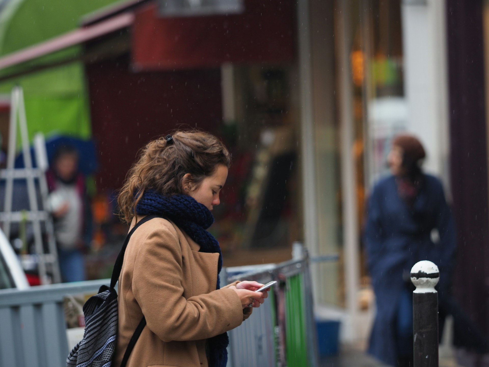 A woman walking outdoors on a rainy day, looking down at her phone while wearing a tan coat, a dark scarf, and a backpack.