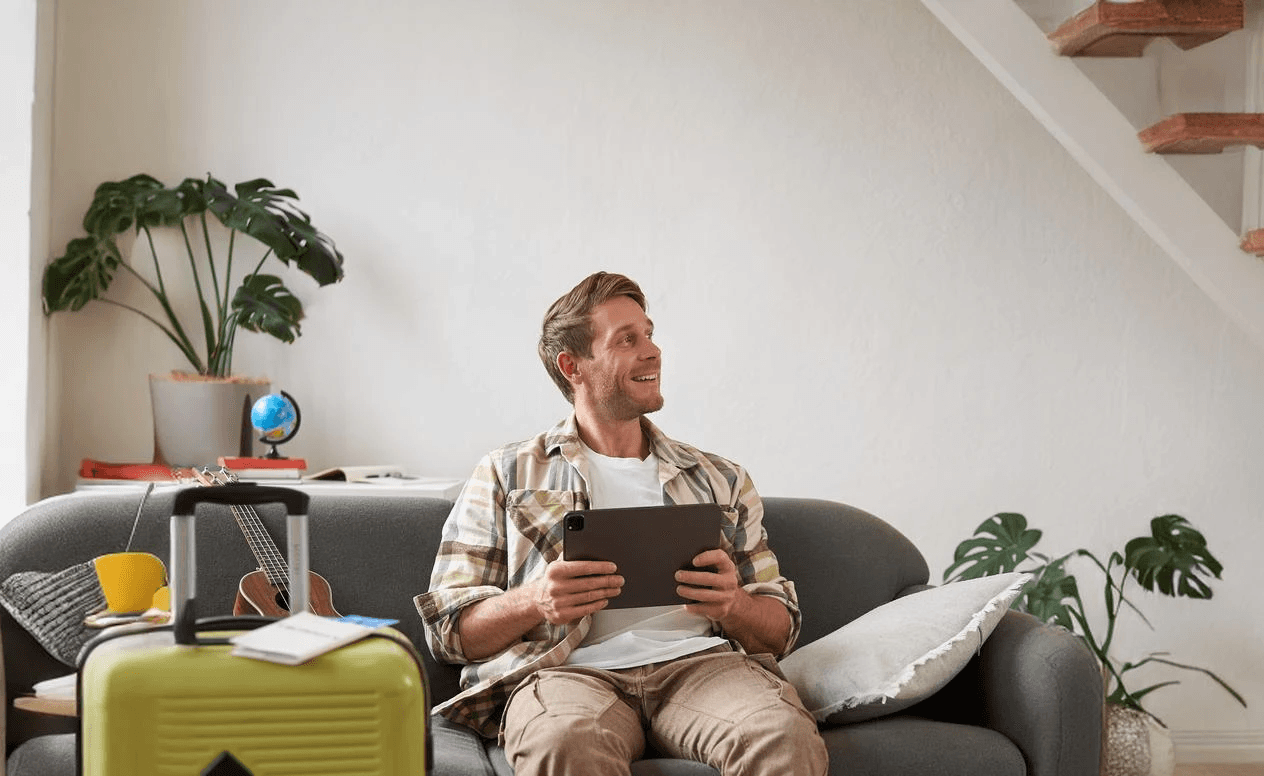 Man sitting on a couch with a tablet and suitcase in a cozy living room.