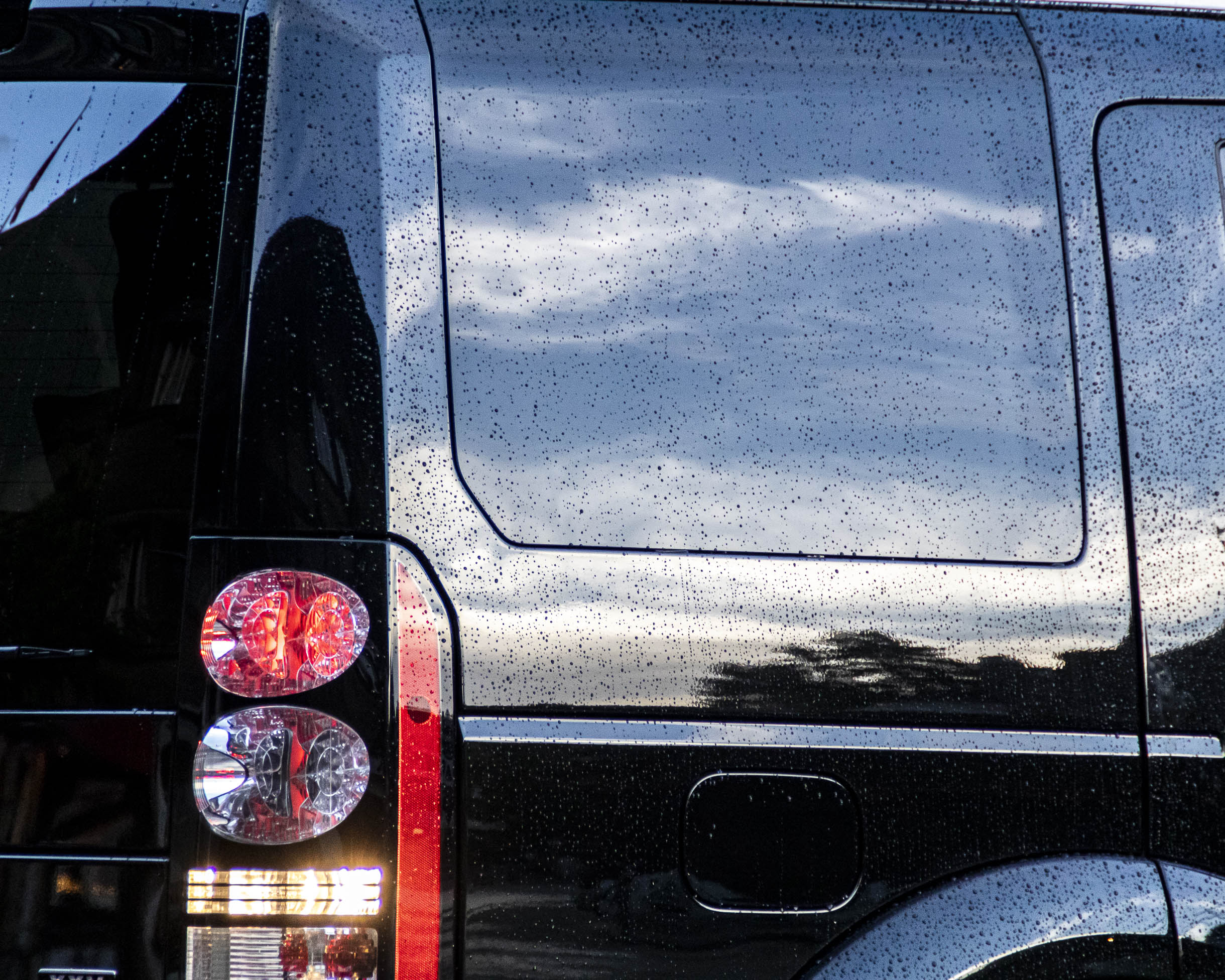 Gros plans sur la carrosserie mouillée d'une Range Rover noir dont le ciel se reflète dans la pleinture