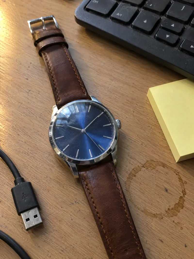 Beispiel-Ausgangsbild, mit KI erstellt: Armbanduhr mit blauem Zifferblatt und braunem Lederband auf einem Bürotisch neben der Tastatur