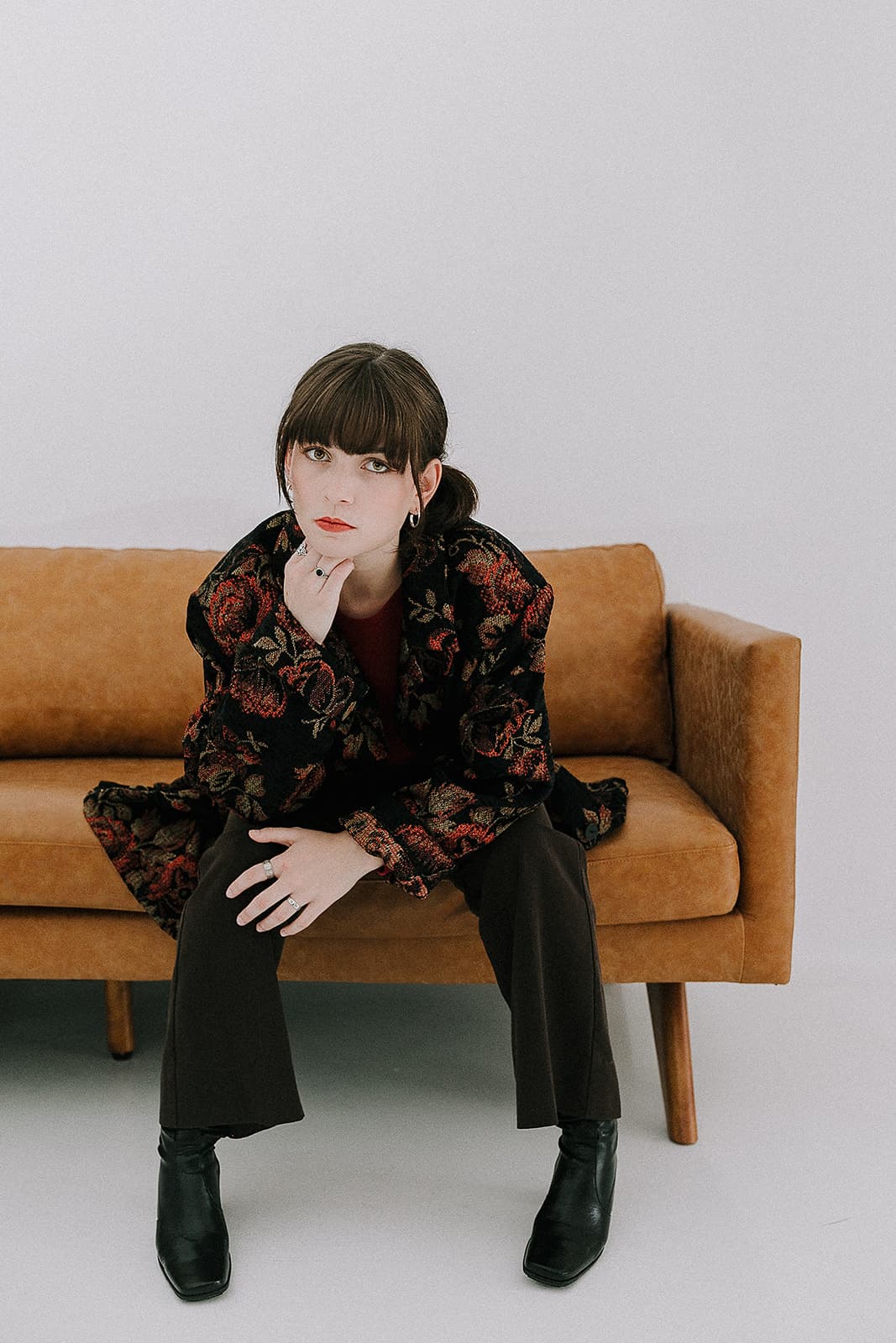 A model leaning forward on a tan couch, wearing a floral-patterned jacket and black boots, embodying the 90s aesthetic at Revelator Studio in Shreveport.