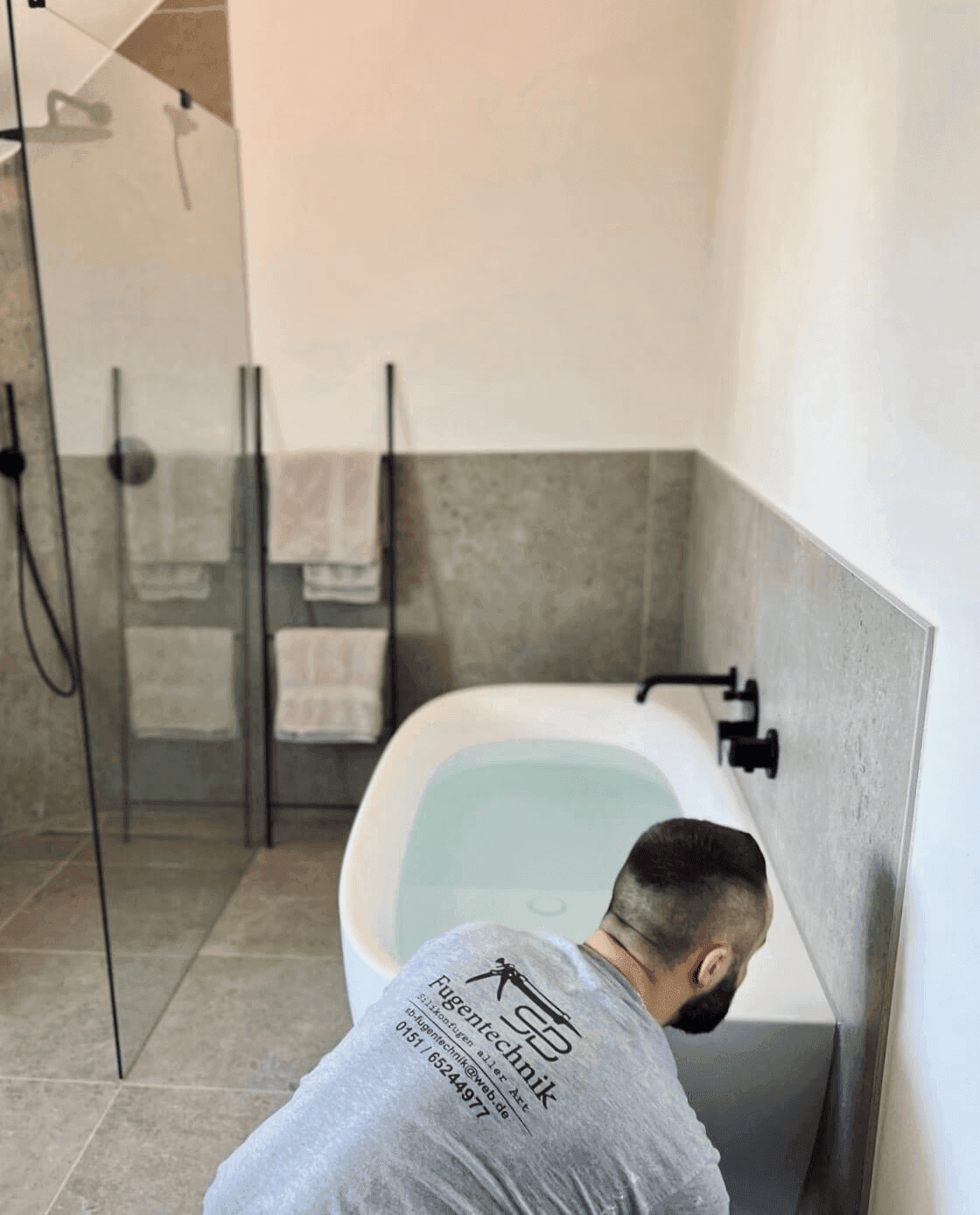A person is seen cleaning or inspecting a bathtub in a modern bathroom with light walls and minimal decor.