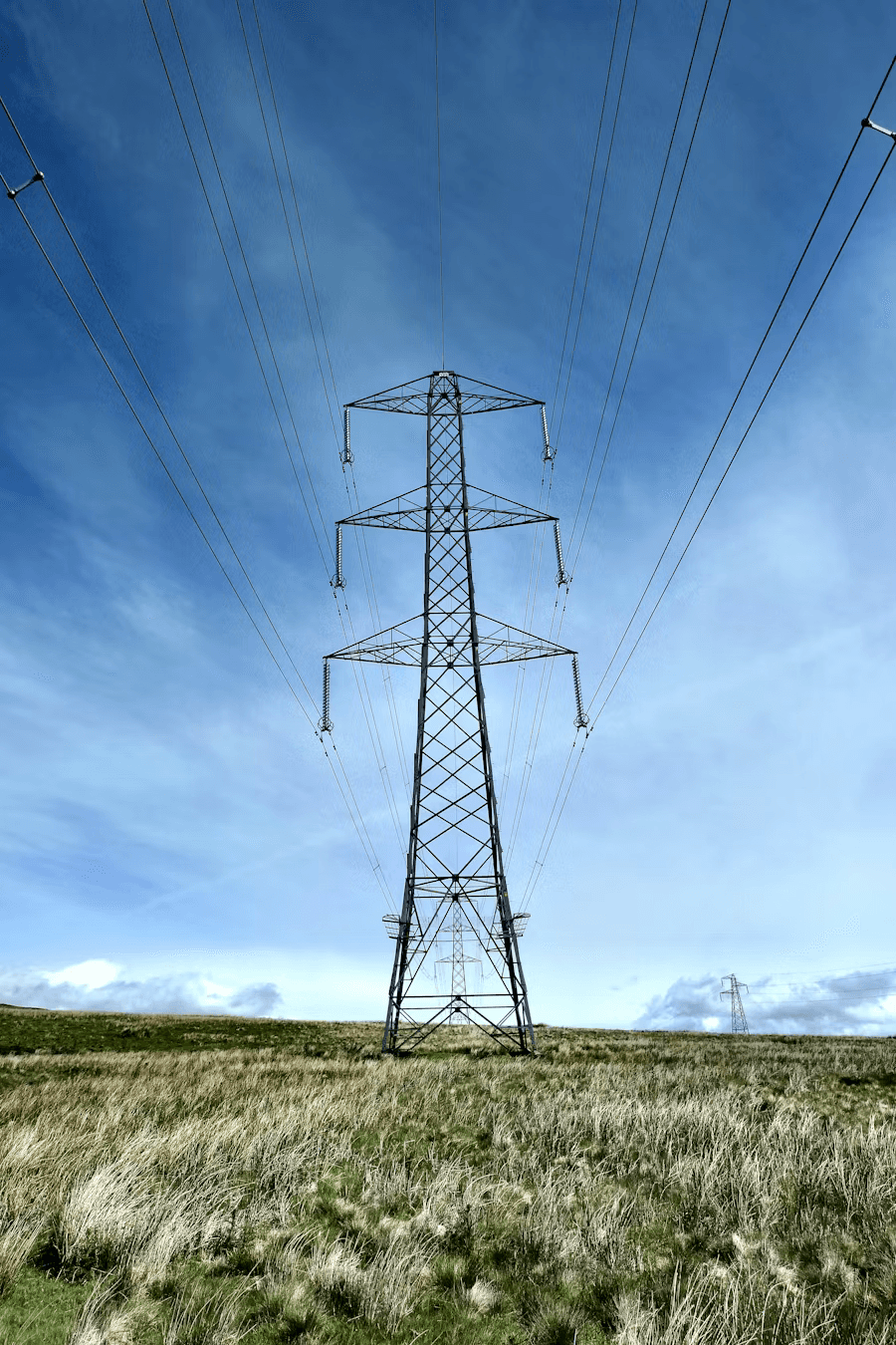 High-voltage electricity transmission tower standing in an open field under a blue sky.