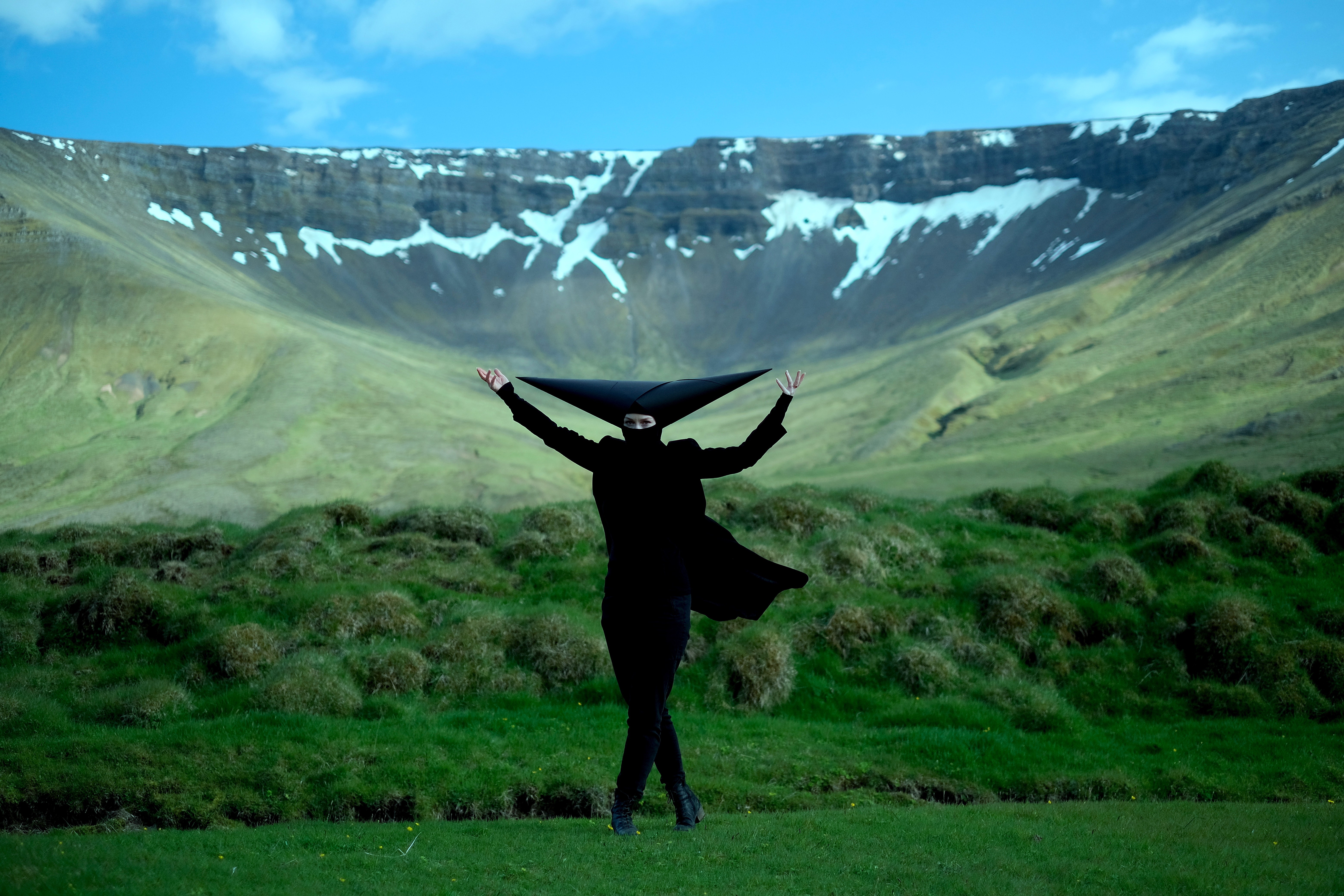 A still from Munnhola, obol ombla obla by Ásta Fanney Sigurðardóttir: a figure dressed in black with a tall pointed headpiece, arms raised, on green moss before a long, snow-streaked Icelandic escarpment.