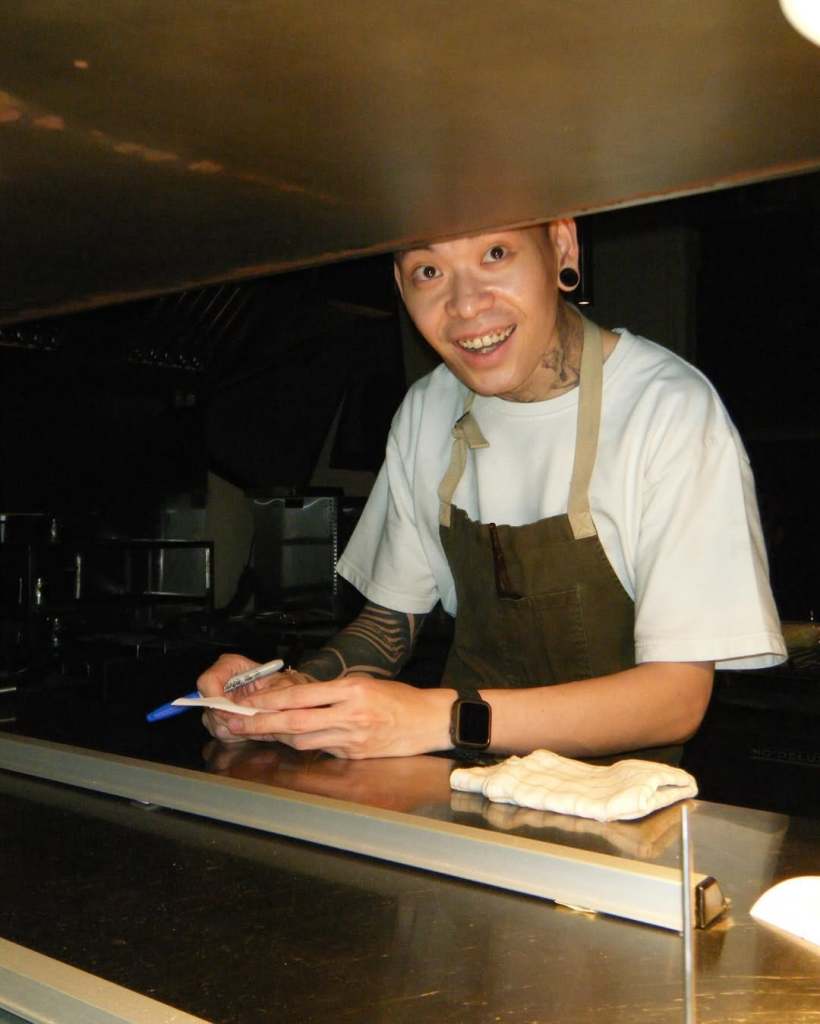 A chef in an apron smiling while writing on a notepad in a kitchen, with a dimly lit background.