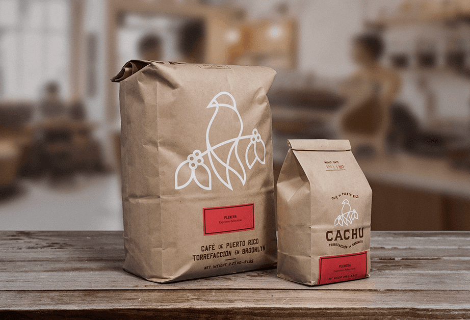 Two brown paper bags sit on a wooden surface. The larger bag on the left has a white line drawing of a bird perched on a branch with three circles. The smaller bag on the right displays the word "CACHU" along with a similar bird drawing and the words "CAFE DE PUERTO RICO" and "TORREFACIÓN EN BROOKLYN." Both bags have red rectangular labels at the bottom. The background is blurred, showing a kitchen or cafe setting.