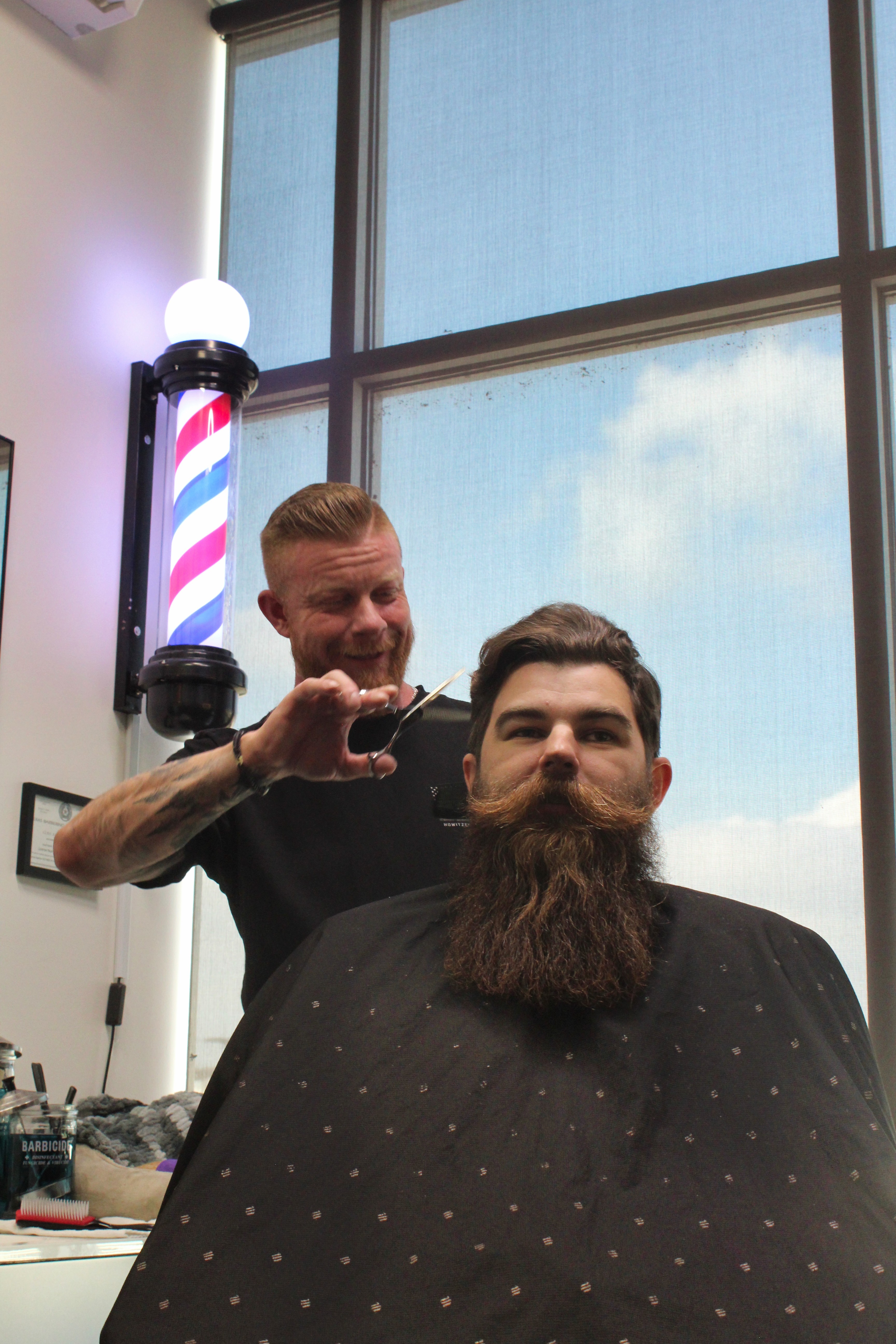 Master barber Matt Finner trims a client’s beard at Minnesota Matt’s Barbershop in Euless, TX, with a traditional barber pole in the background. Natural daylight fills the room, highlighting the relaxed and professional grooming experience.