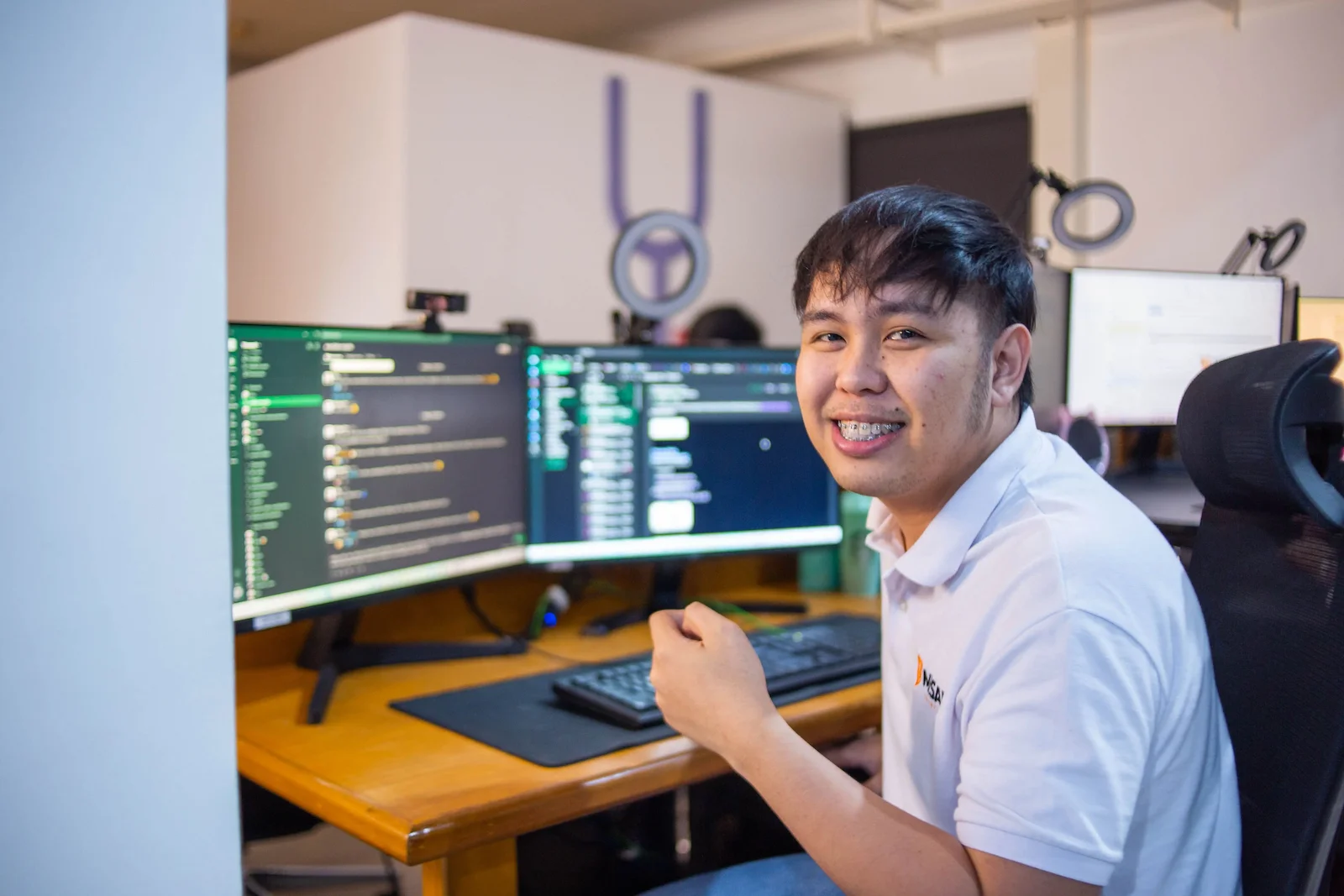 A young man in a white shirt looks up from his computer with a smile, seated at a desk with code on the screen.