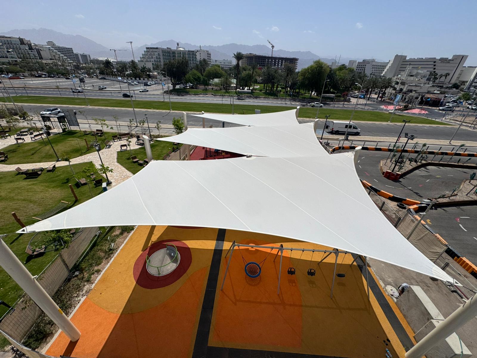 Playground with swings under large beige sunshades next to a road and buildings in the background.