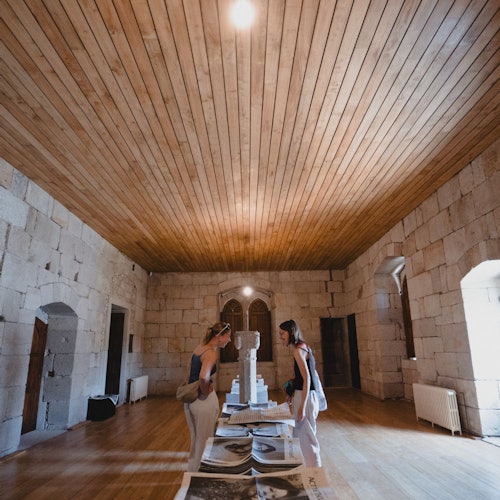 两个人在一个石墙的房间里，天花板是木制的，正在查看中央桌上的展览物品。