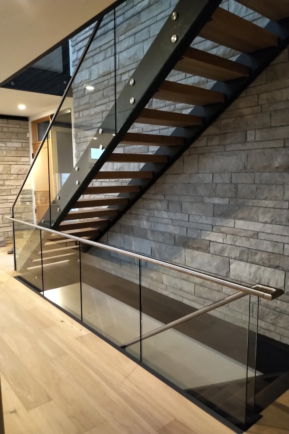 Modern glass stair railing installed on a steel stringer staircase with wood treads and stone feature wall.