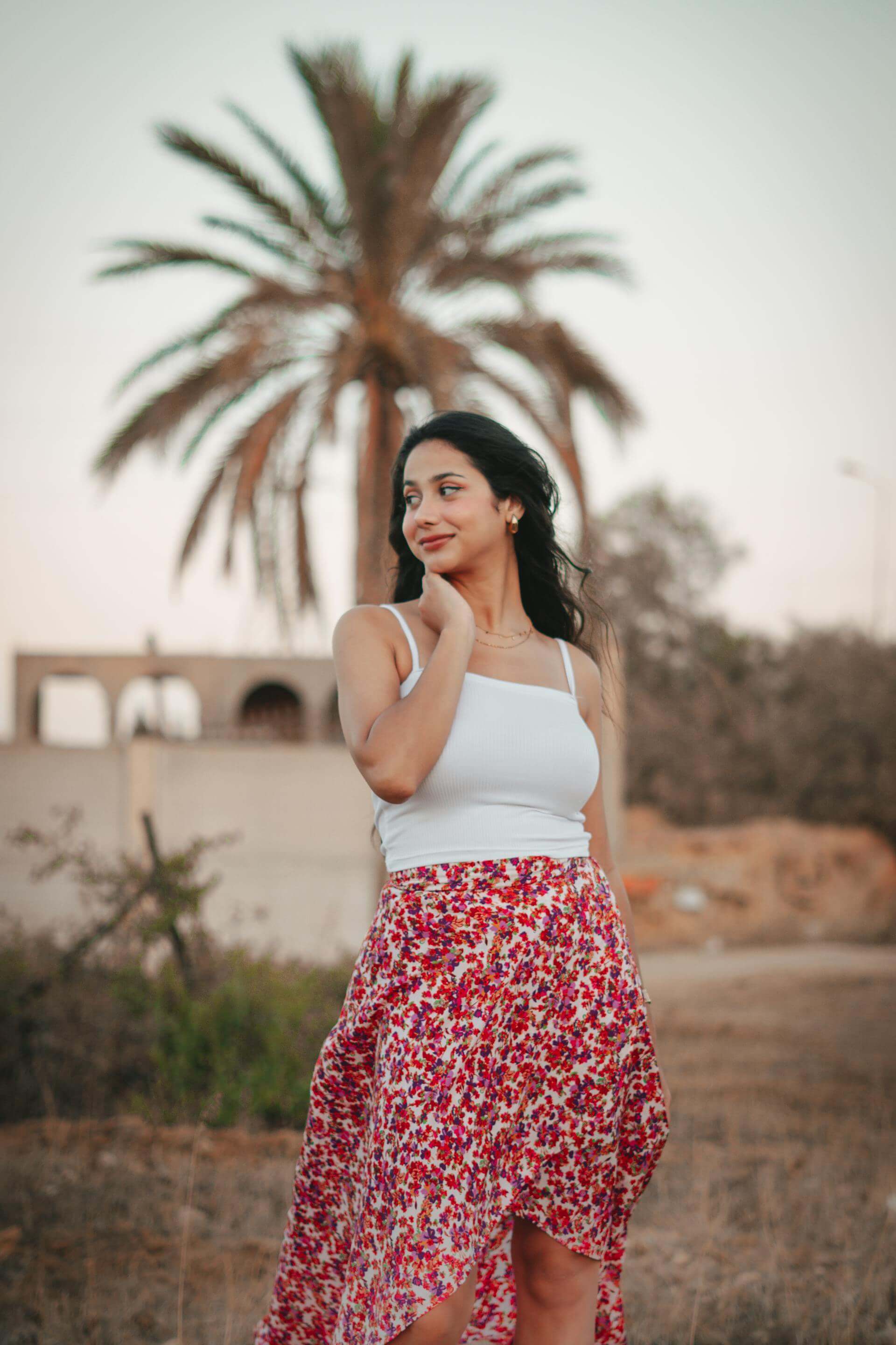 A woman wearing a white tank top and a red floral skirt stands confidently, showcasing her vibrant outfit