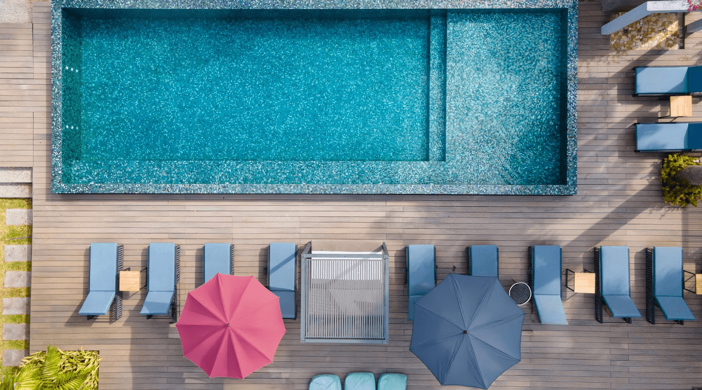 Aerial view of a modern rectangular swimming pool with surrounding timber deck, sun loungers, and colourful umbrellas.
